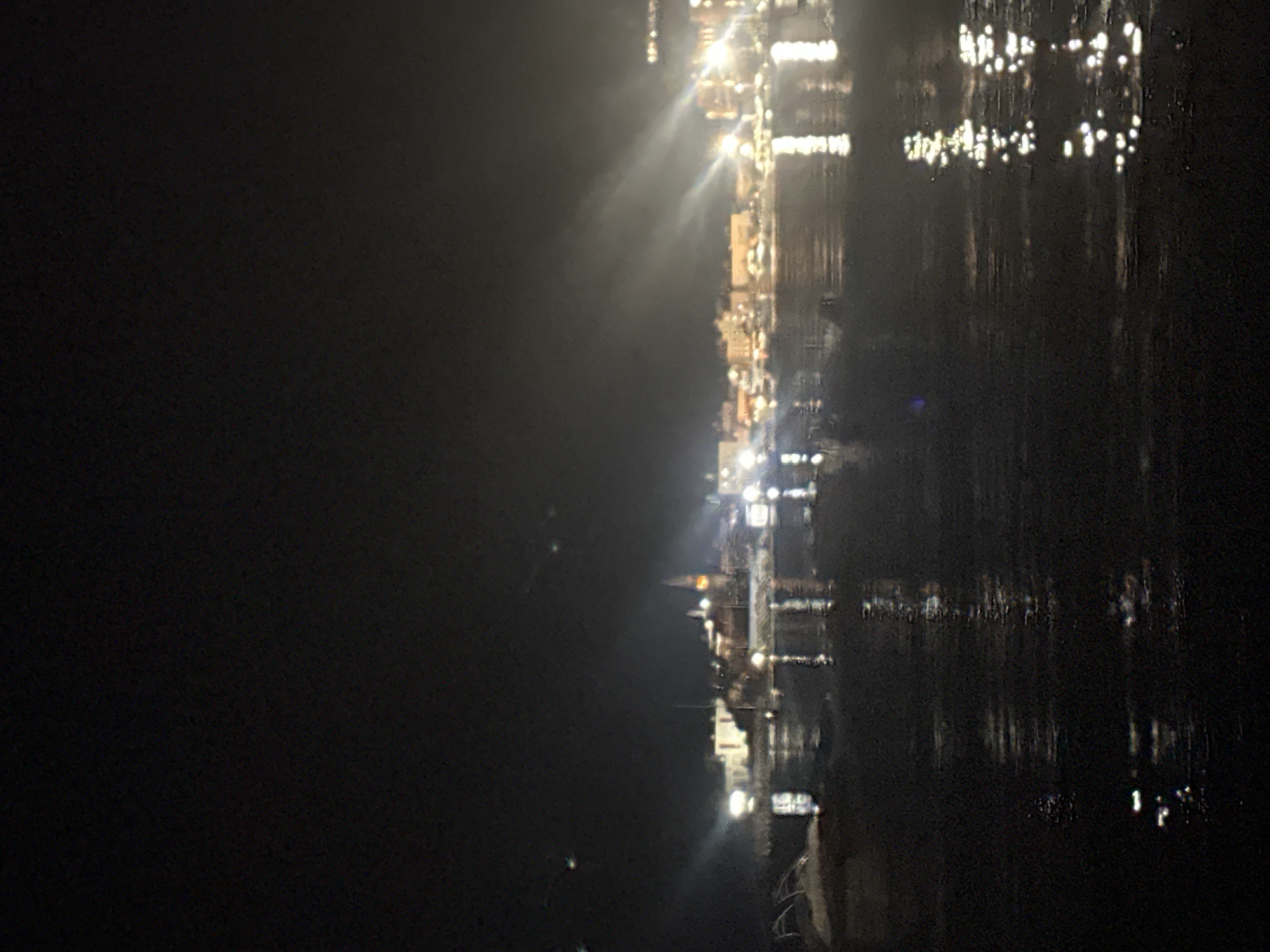 The image presents a nighttime scene overlooking the coastal city of Budva, Montenegro, likely from across the bay or from a higher vantage point. In the foreground, a dark body of water, possibly the Adriatic Sea, reflects numerous bright, elongated lights from the distant shore, creating shimmering vertical streaks on its surface. The midground and background feature a densely illuminated urban landscape. On the left, distinctive historic architecture, including a prominent church tower or spire, is brightly lit, corresponding to Budva's Old Town. To the right, a wider expanse of buildings, also densely illuminated, forms the remainder of the cityscape. Several strong beams of light emanate upwards and outwards from various points within the town, cutting through the dark night sky, suggesting the presence of atmospheric haze or fog. No people or specific actions are discernible within the frame. The scene is characterized by the stark contrast between the luminous city lights and the surrounding darkness of the night and water.