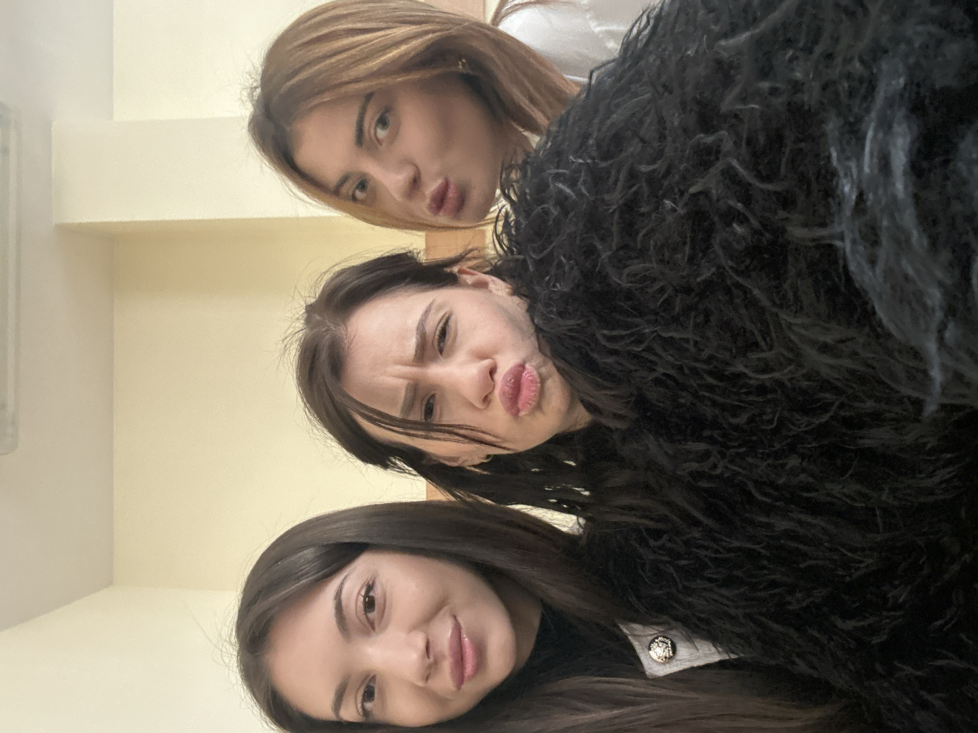 Three young women are arranged in a stacked pose, facing the camera with playful expressions. The woman at the top has light brown hair and is puckering her lips. The woman in the middle has dark brown hair, pulled back, and is also puckering her lips while raising her eyebrows. The woman at the bottom has long dark brown hair and is smiling slightly. A large, dark, shaggy garment is draped over the middle and bottom women, obscuring much of their clothing. The background is a plain wall painted in a light beige color. The location is likely indoors. There is no immediately discernible indication the photograph was taken in Plovdiv, Bulgaria.