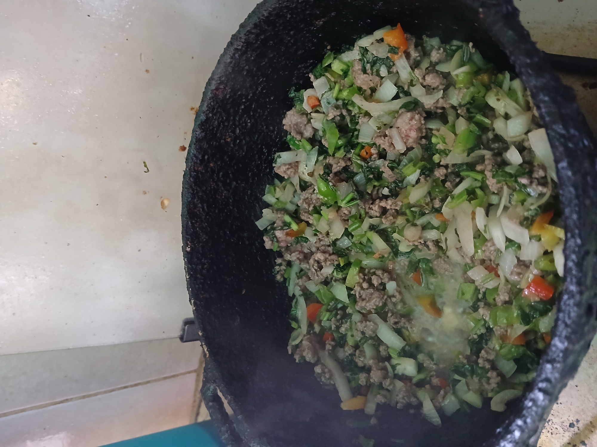 The image shows a cast iron pot filled with a mixture of cooked ground meat and various chopped vegetables, including onions, green peppers, and other colorful vegetables. The pot appears to be resting on a stove or cooking surface, with a white tiled wall visible in the background. The location of the image is mentioned as the city of Juan Griego, Venezuela.