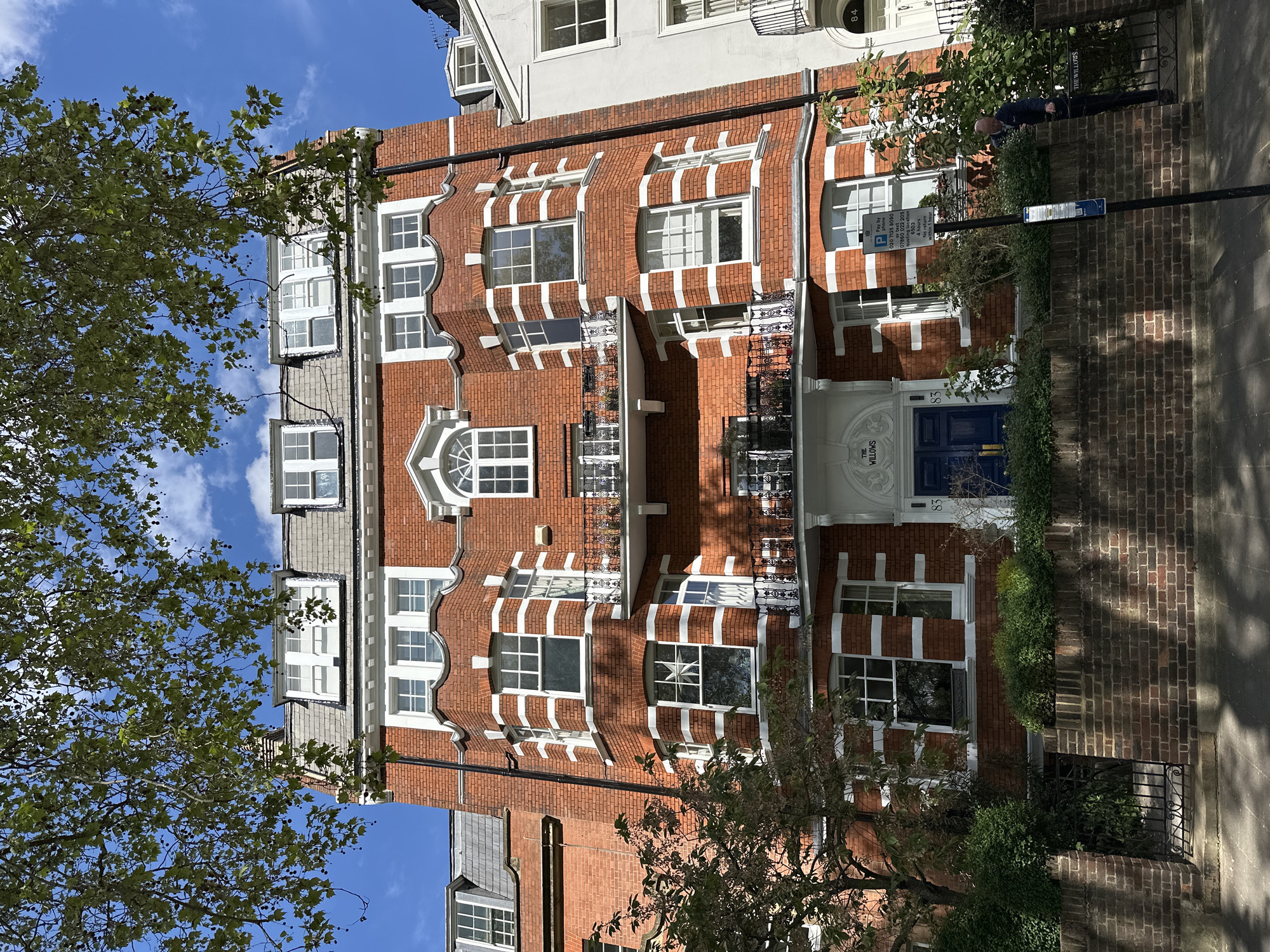 The image shows the exterior of a red brick building in London, United Kingdom. The building has white window frames and accents. A blue door is visible with the name "The Willows" above it. A parking sign is present, indicating parking restrictions. The building's architectural style appears to be somewhat historic. Trees partially obscure the view of the building. A portion of the adjacent building is also visible.