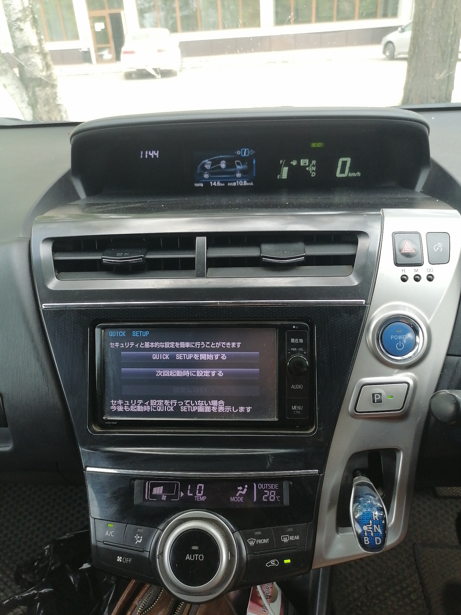 The image shows the interior of a car, specifically the dashboard and center console. A digital display in the upper part of the dashboard shows the time as "11:44" and other vehicle information including speed and average fuel consumption. Below this, a touchscreen display presents menu options in Japanese, with one option highlighted. To the right of the touchscreen are various buttons, including a power button and a parking brake release. The center console features climate control buttons and a gear shifter. The car appears to be parked. The provided context states the location is Lesozavodsk, Russia.