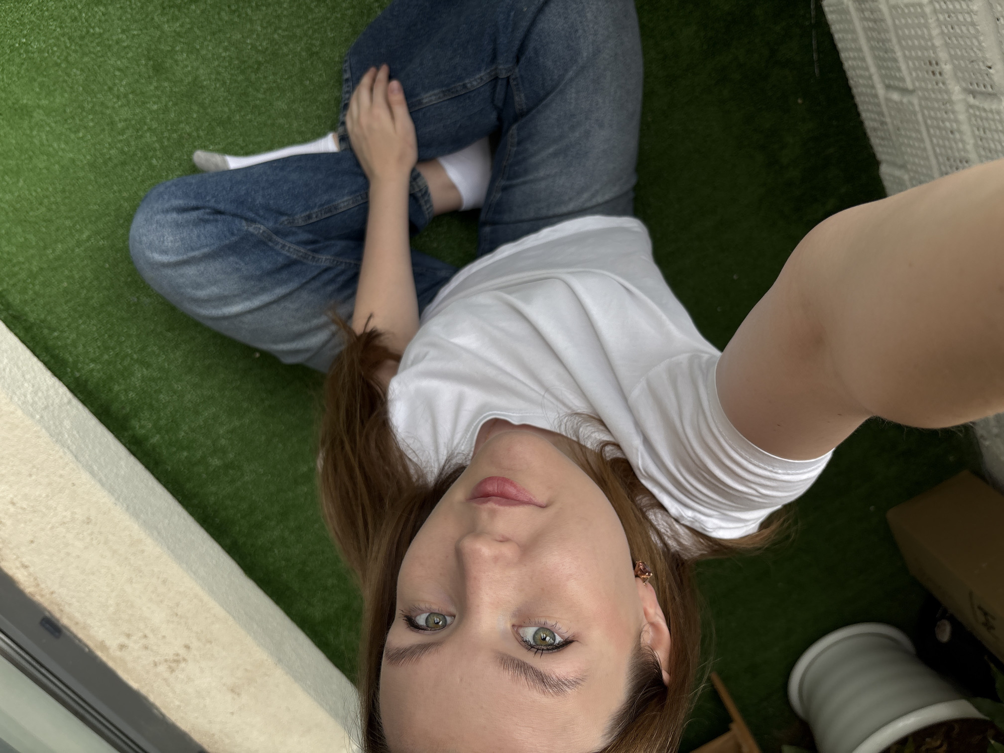 A young woman with long brown hair is sitting on an outdoor balcony with artificial turf. She is wearing a white t-shirt and blue jeans, and white socks. She is looking at the camera and holding the phone to take a selfie. The balcony has a white railing and a beige wall. There is a white pot with a plant on the left side of the image, and a cardboard box behind it. The image does not provide any specific clues to determine the location in Izhevsk, Russia.