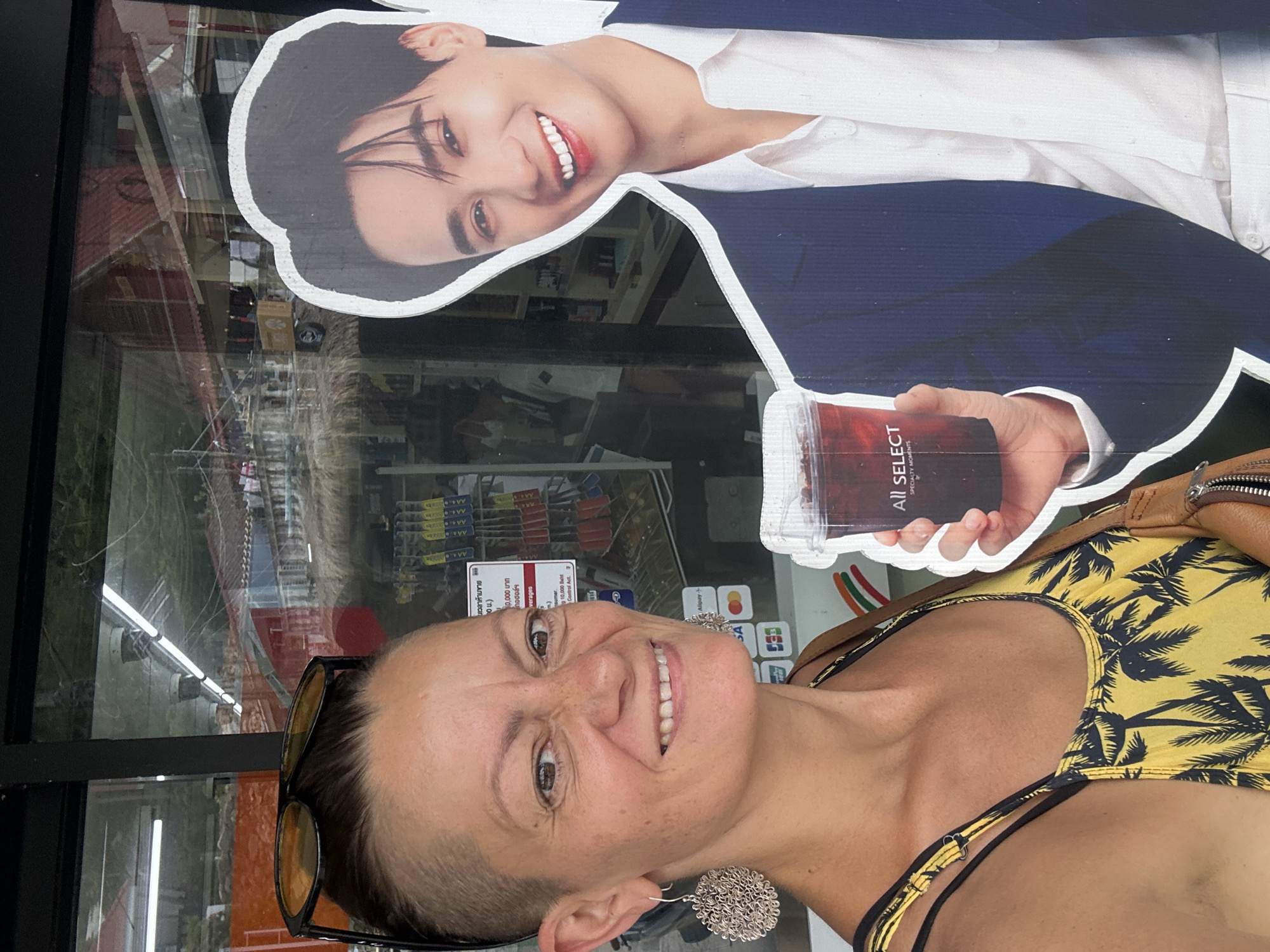 A woman with a shaved hairstyle, wearing sunglasses and a yellow and black palm-print dress, smiles at the camera. She is standing next to a life-sized cardboard cutout of a smiling man in a blue suit and white shirt. The man's cutout is holding a clear cup with a dark red beverage. They are in front of a store with a large glass window displaying shelves of products. The background through the window shows more shelves, possibly of food and drinks, and a glimpse of the outside with a thatched roof structure. The scene appears to be on a street in a tropical location.