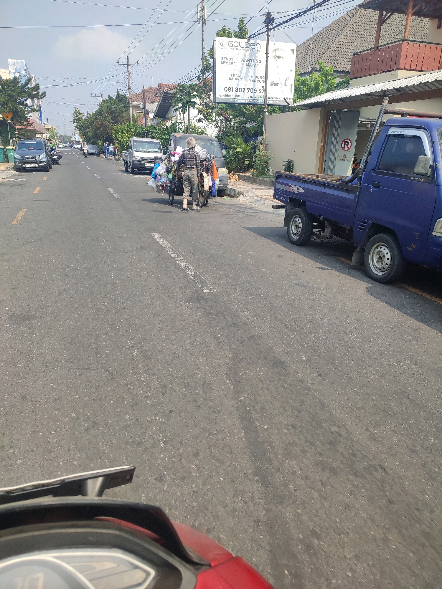 The image depicts a street scene in Yogyakarta, Indonesia, likely on a sunny day. In the foreground, the handlebar and dashboard of a red motorcycle are visible, indicating the photo is taken from a rider's perspective.
The street is paved and appears to be a main thoroughfare, with vehicles parked along the side and moving in the same direction. On the left side of the road, a black car with license plate "AB 1696 UT" is parked. Further down the street, several other cars and motorcycles are visible.
A significant element of the scene is a person, likely a street vendor, pushing a cart laden with various items, including plastic bags and what appears to be recycling materials. This vendor is walking in the middle lane of the road. Next to them is a silver truck.
On the right side of the road, a blue pickup truck is parked. Behind the truck, there is a building with a tiled roof and an overhang. A large billboard is mounted on a pole on the right side of the street, featuring text in Indonesian and an image of an eye. The text mentions "GOLDEN AESTHETICS AND DERMATOLOGY CLINIC" and provides a phone number "081 802 709 371".
The environment suggests a typical urban street with a mix of residential and commercial buildings. Power lines and utility poles are visible overhead, indicating the presence of infrastructure. The sky is light blue with some clouds. The overall impression is that of an active street in a populated area of Yogyakarta.