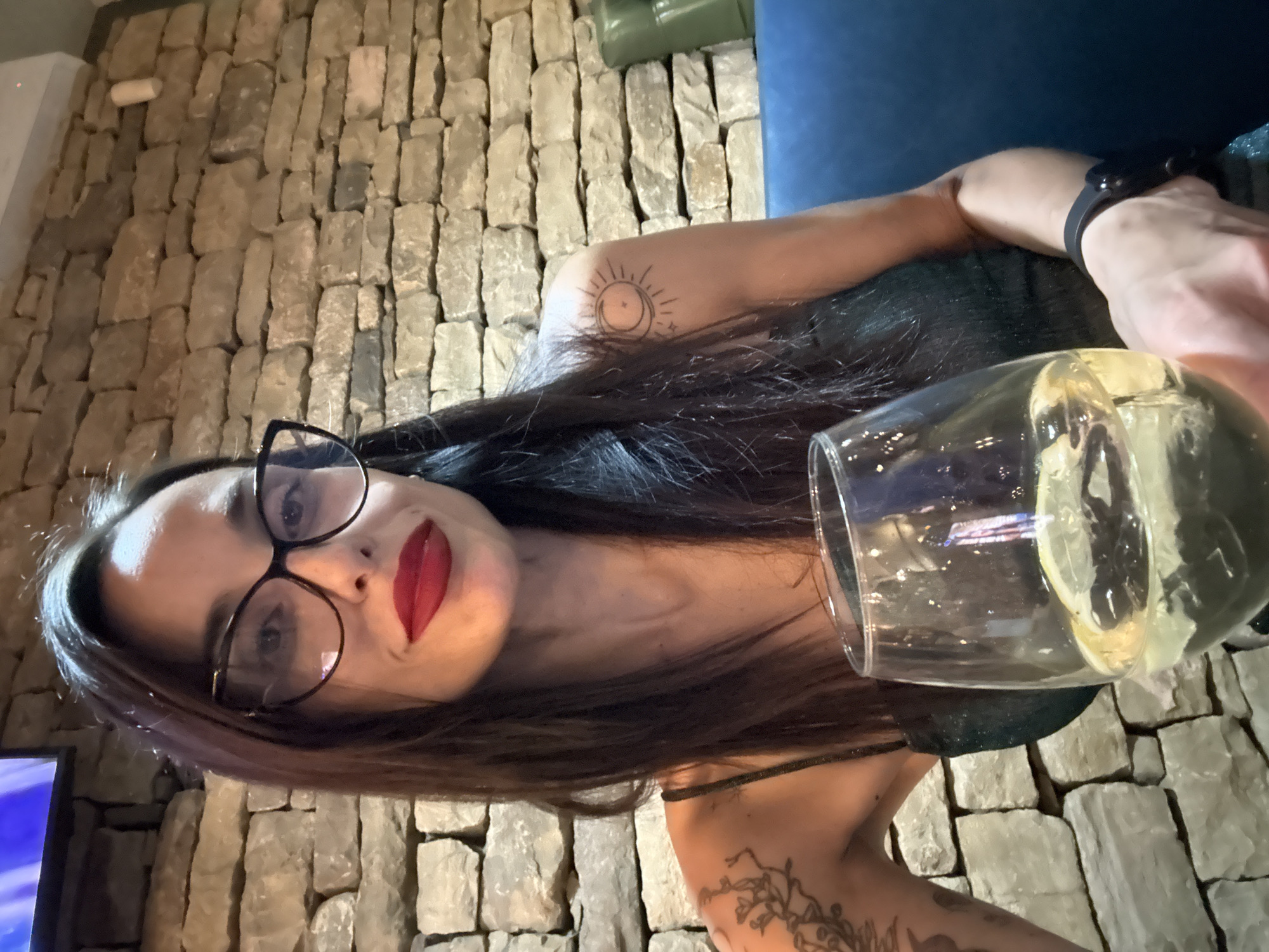 A woman is holding a glass of liquid while facing the camera. She is wearing glasses and red lipstick, and has tattoos on her arms. The background is a brick wall. Based on the location and the available details, the scene might be indoors at a bar or restaurant in Pleven, Bulgaria.