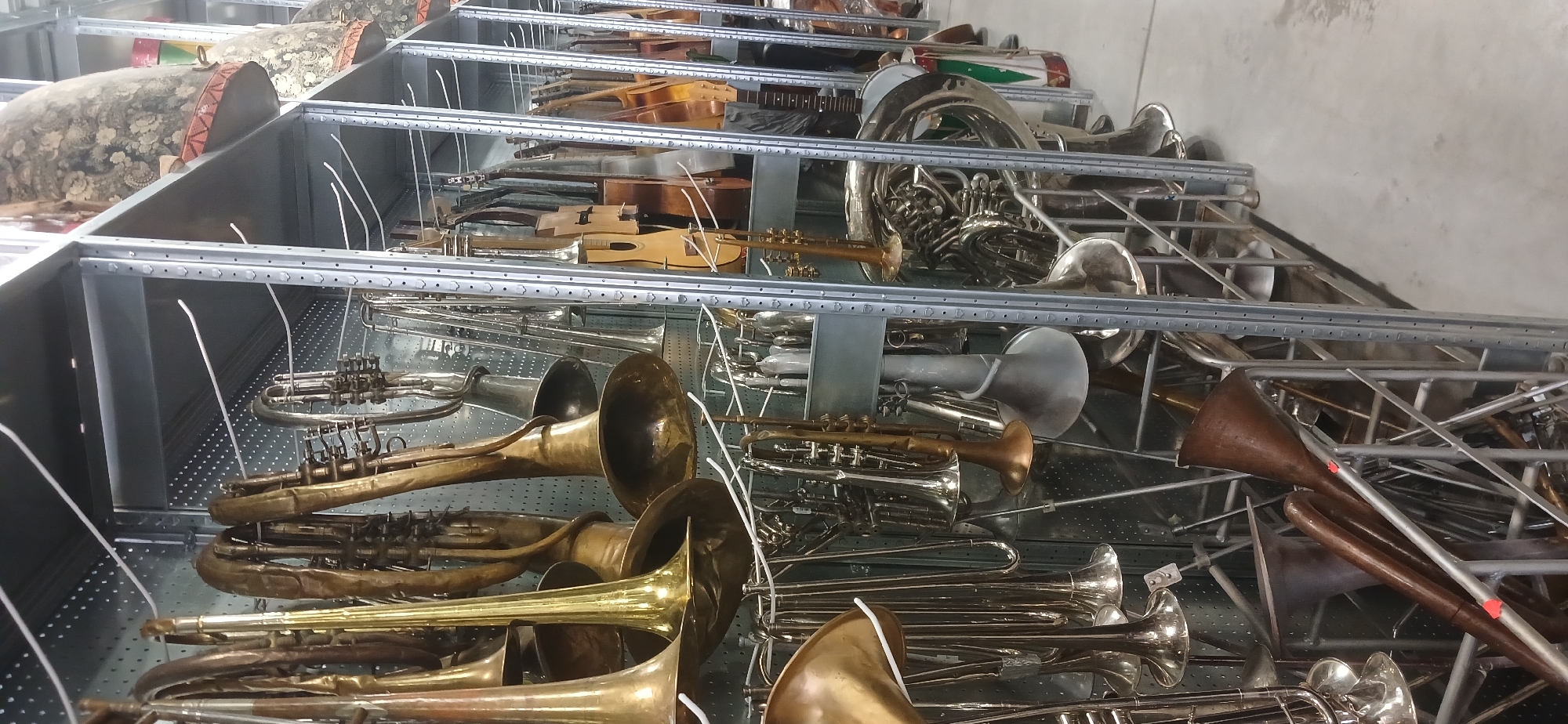 This image shows a storage area filled with various musical instruments. The instruments are stored on multiple levels of a metal shelving system, seemingly constructed from perforated metal sheets. The instruments include a variety of brass instruments such as trumpets, trombones, tubas, and potentially French horns. There also appear to be stringed instruments such as guitars and possibly mandolins. Drums of various sizes are also visible. The storage area seems to be indoors, possibly in a warehouse or large storage facility. The specific location and city are unknown.