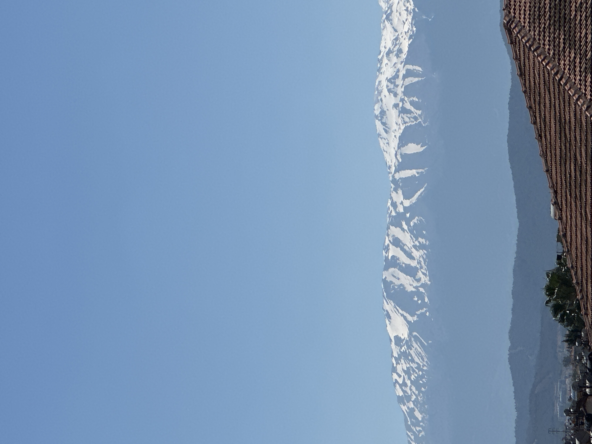 The image shows a sideways view of snow-capped mountains taken in Fethiye, Turkey. The sky is clear blue, and to the right of the mountain range, the edge of a building roof is visible. At the base of the mountains, sparse trees and the tops of buildings can be seen.