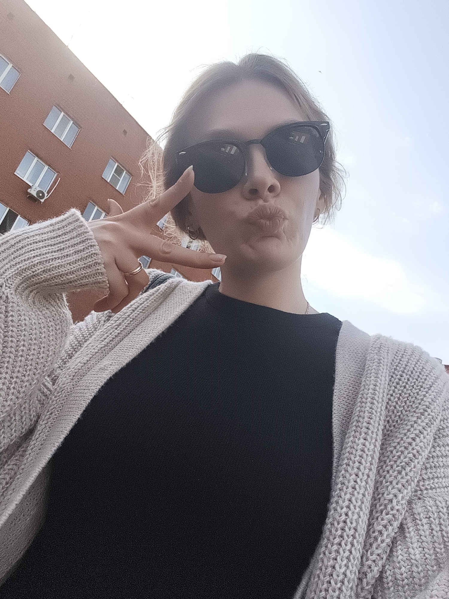 The image shows a woman taking a selfie outdoors. She is wearing sunglasses, a black top, and a light gray knitted cardigan. She is making a peace sign with her left hand and puckering her lips in a kissing gesture. Behind her is a brick building with windows and an air conditioning unit. The sky is visible in the upper portion of the frame. Based on the architectural style and the building design, the location is likely in a city in Russia, potentially Izhevsk, Russia.