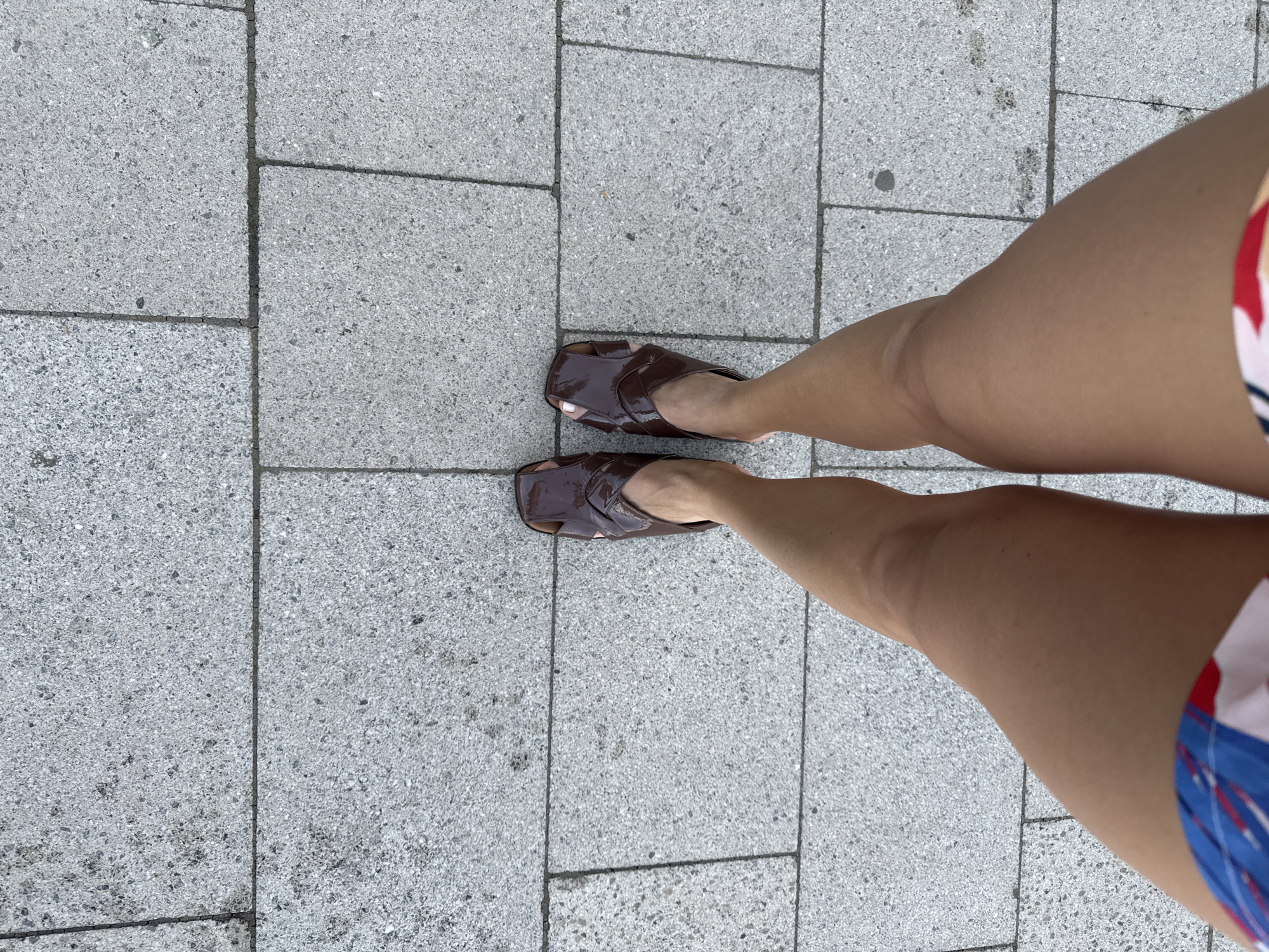 The image presents a view of a person's legs and feet standing on a paved surface, likely a sidewalk or pedestrian area. The paving consists of rectangular, light gray stones. The person is wearing brown, patent-leather-like sandals and shorts with a pattern of red, white, and blue. The photograph is taken from directly above, providing a close-up perspective of the lower body and the ground immediately surrounding it. The setting is urban, suggested by the paving stones and the overall clarity of the image. Based on the prompt, the location is in the city of Chaikovsky, Russia.