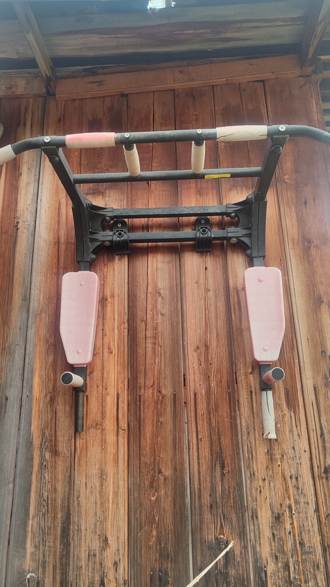 A pull-up bar is mounted on a weathered wooden wall. The pull-up bar is black metal with several grips. Two of the grips are light pink and cracked, while others are white and cylindrical. Below the main bar, there are two pink padded arm rests with cylindrical handles extending downwards. The wooden wall is made of vertical planks of varying shades of brown, showing signs of age and weathering. The scene appears to be outdoors, possibly on a shed or an exterior wall. The location is specified as Mirny, Russia. There are no people or animals present in the image.
