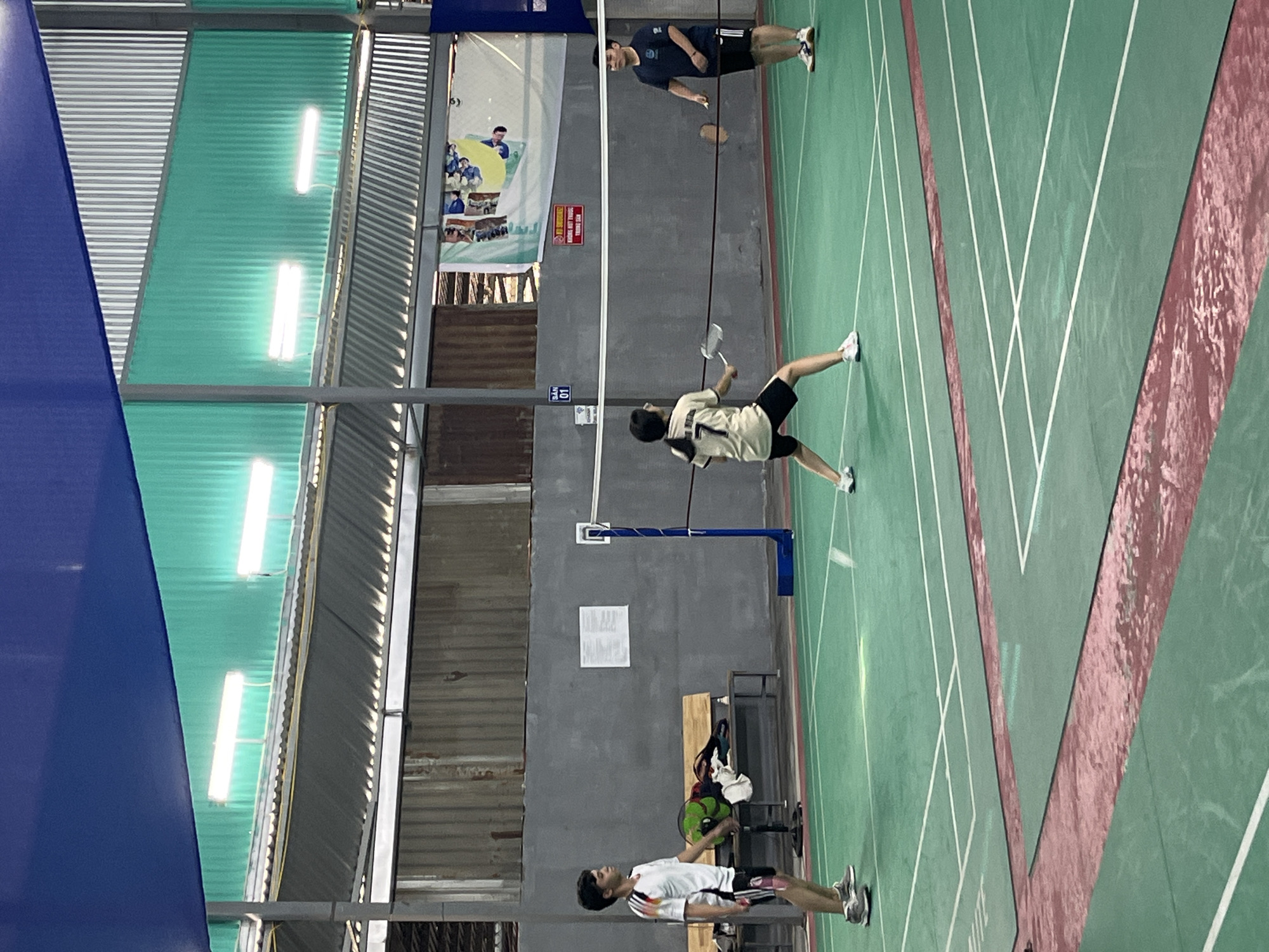 The image depicts an indoor badminton court with a green surface marked with white lines. Three individuals are present: a young male in a white and black shirt and shorts on the left, a young female in a beige and black outfit with the number "7" on her back in the center, and a male in a navy blue shirt and black shorts on the right. The young male on the left is standing, facing towards the center of the court. The young female in the center is in a playing stance, holding a badminton racket, and appears to be mid-game, facing the male on the right. The male on the right is also in a playing stance, holding a racket, and facing the female. The court is enclosed by grey walls and has a metal roof structure above, with blue and green corrugated panels and fluorescent lighting fixtures. A badminton net is strung across the center of the court. In the background, there are benches, sports equipment, and a banner with images. The overall scene suggests a casual game of badminton taking place in an indoor sports facility.