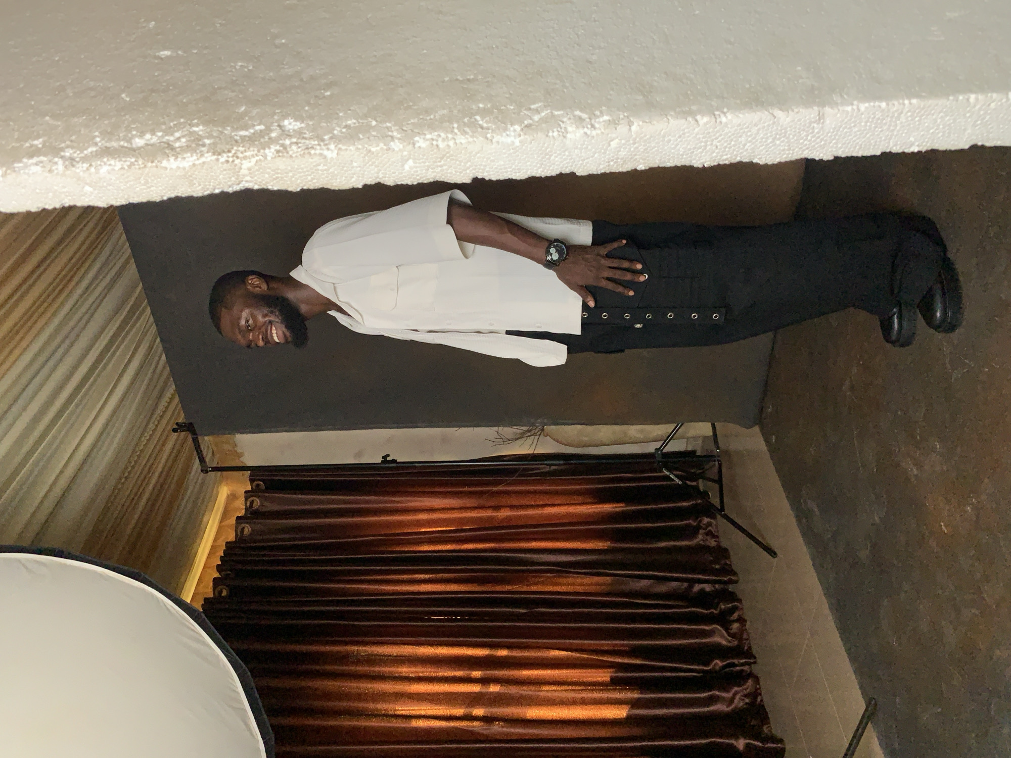 A man with a beard, wearing a white short-sleeved shirt and black pants, stands on a tiled floor. He is smiling and looking to his left. A watch is visible on his left wrist. Behind him, a dark brown curtain is draped. To the left of the curtain, a white circular object, possibly a softbox or umbrella used in photography, is visible. The ceiling above appears to be made of a white, textured material. The overall scene suggests an indoor setting, possibly a studio or a room with photography equipment. The image is oriented vertically, with the top of the frame appearing at the left.