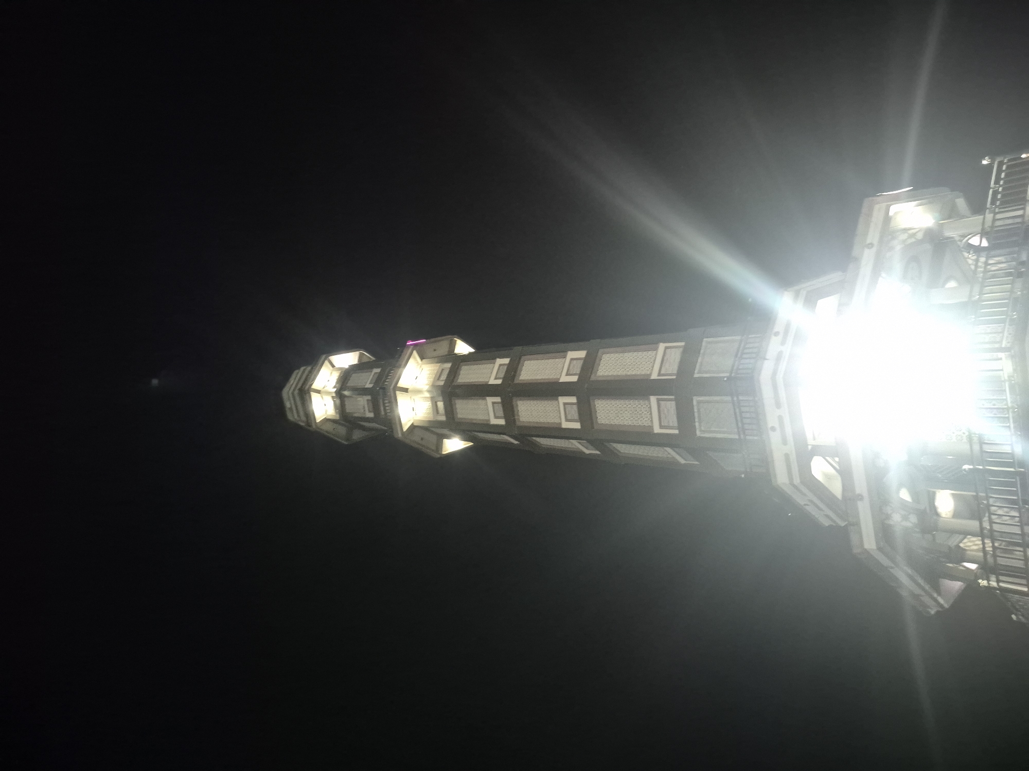 A low-angle, nighttime photograph depicts a tall minaret structure in Keudekruenggeukueh, Indonesia, brightly illuminated against a dark sky. The minaret has a polygonal shape and tiered construction, with evenly spaced window openings. Bright light emanating from the structure washes out some details. A ladder system is visible on one side of the building. The surrounding environment appears black, indicating night.
