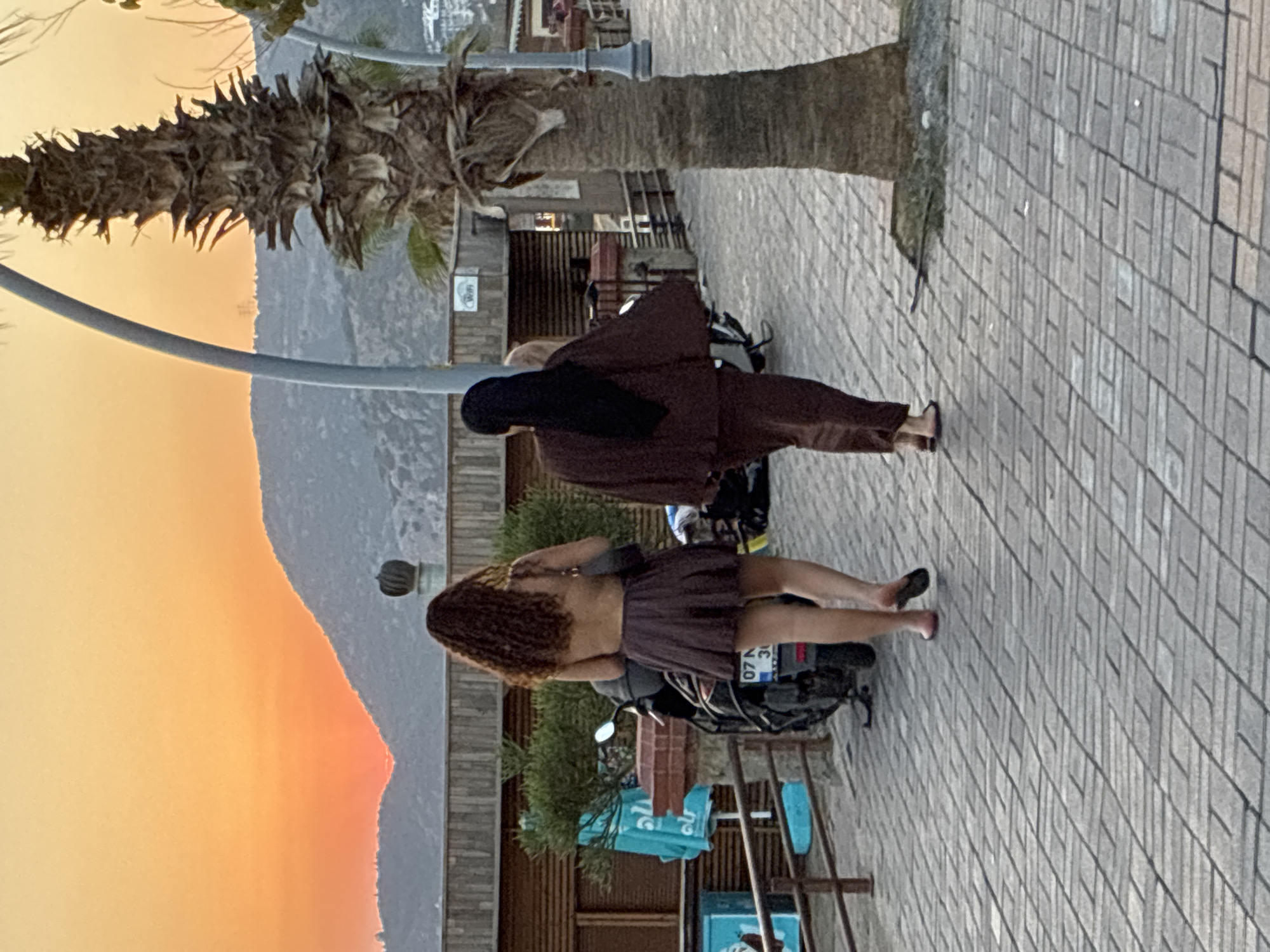 Two women walk away from the camera on a brick-paved promenade in Alanya, Turkey, with a sunset casting an orange glow over distant hills. The woman on the left has long, curly brown hair and wears a low-cut, short brown dress. The woman on the right wears a dark headscarf and a brown outfit consisting of a loose-fitting top and matching pants. A palm tree stands to the right of the frame, and a motorcycle with a license plate is parked to the left. The background shows wooden structures, vegetation, and buildings on a hillside.