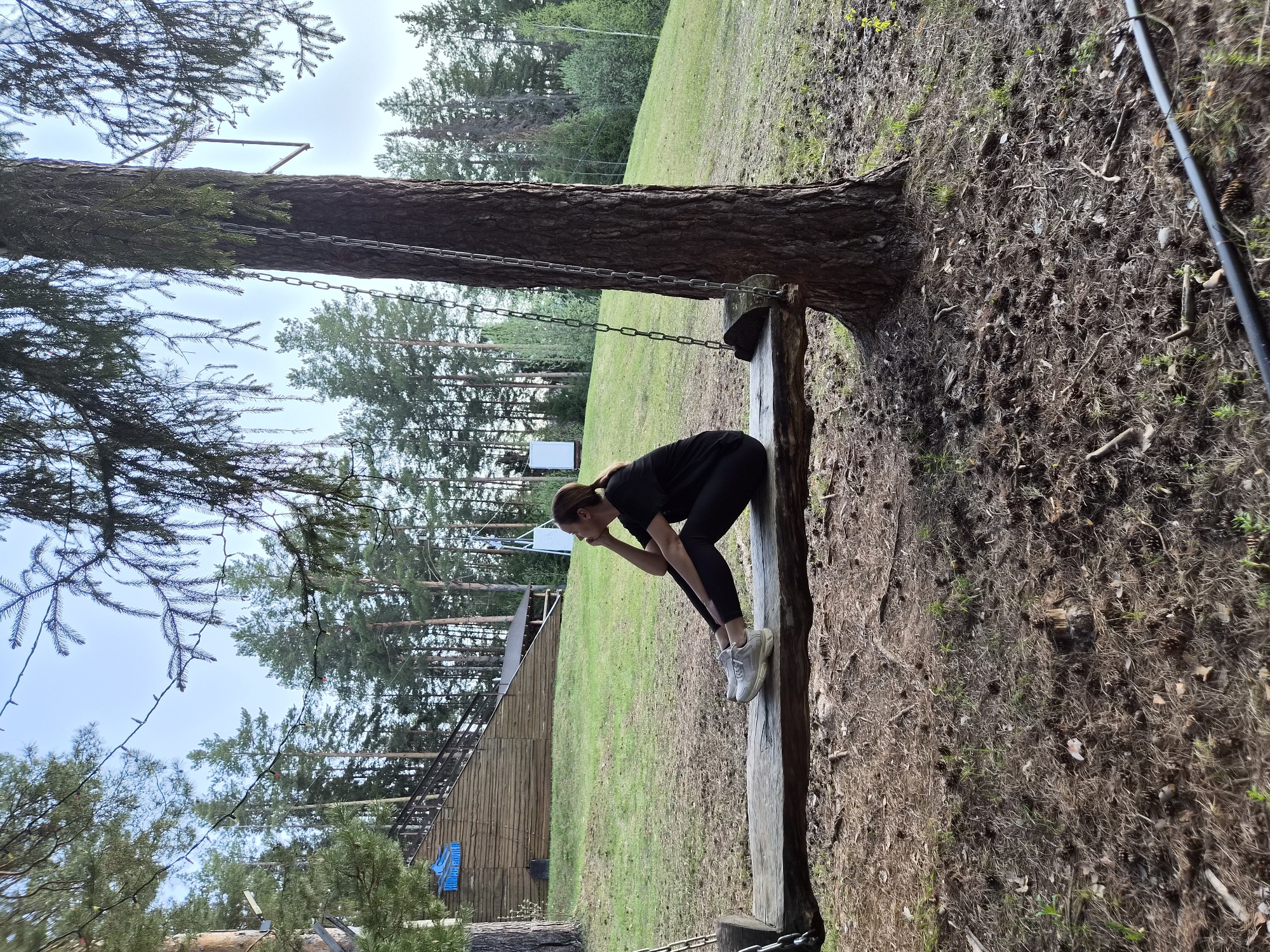 The image shows a woman in black athletic wear standing on a wooden beam in what appears to be a park or outdoor obstacle course in Прикамский, Russia. She is bending forward with her hand on her forehead, seeming to be thinking or taking a break. The beam is attached to a large tree by chains. The ground is covered with fallen pine needles and other debris typical of a wooded area. A wooden structure is visible in the background, along with a grassy area and more trees.