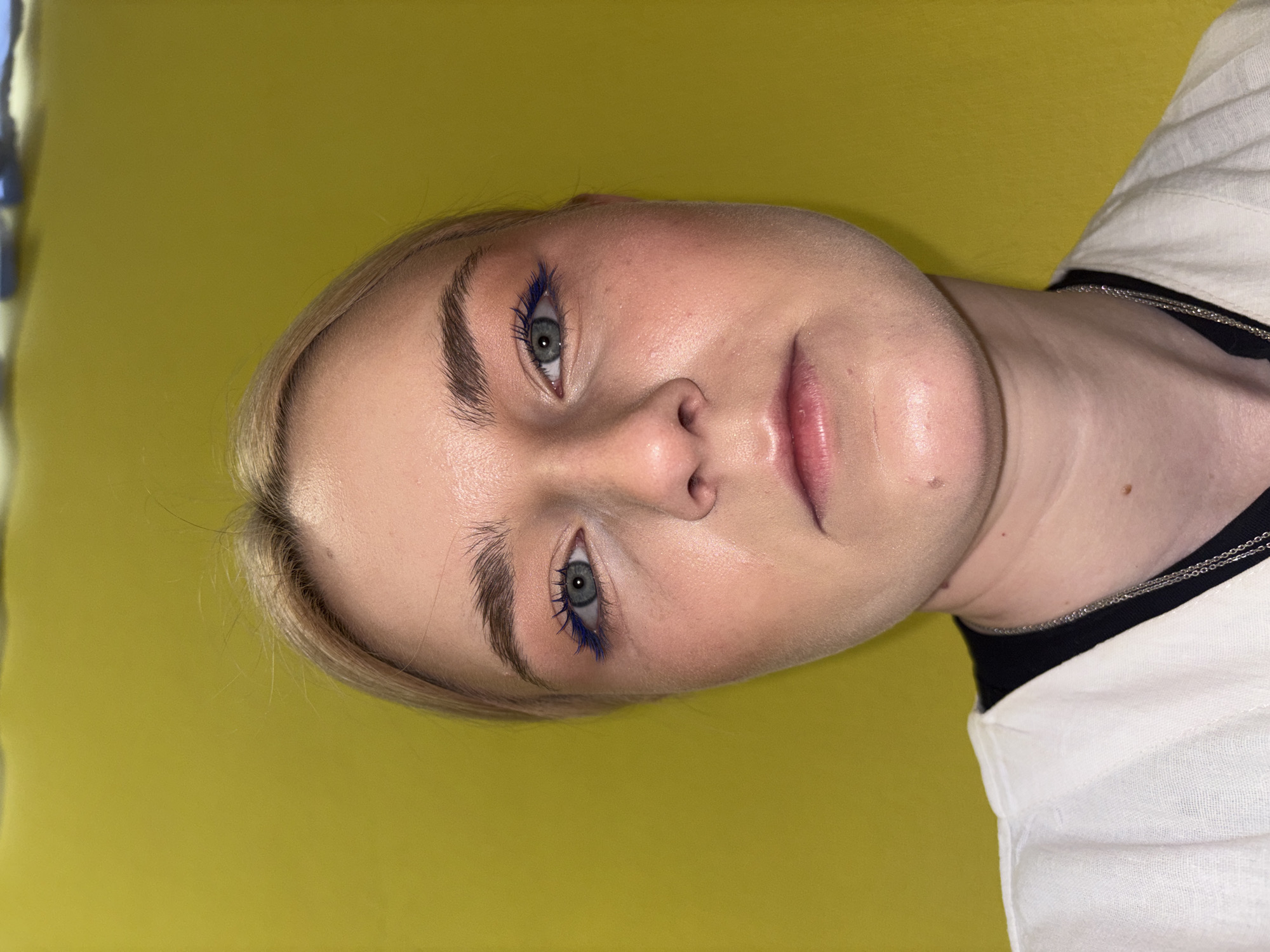 A close-up, head-on, medium shot captures a young woman with fair skin and blonde hair against a vibrant yellow wall. She is wearing a white collared shirt over a black t-shirt, with a silver chain necklace visible. Her makeup includes light foundation, pink blush, defined eyebrows, and bold blue mascara and eyeliner. Her eyes are a light blue-gray, and her lips are bare. The lighting is somewhat harsh, creating a slightly overexposed effect on her face and highlighting the texture of her skin. The background is a solid, textured yellow.