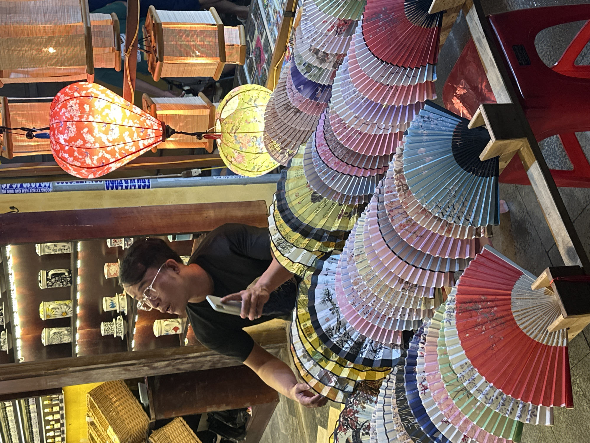 A person wearing glasses and a black t-shirt is standing in what appears to be a market stall in Hội An, Vietnam. They are holding a smartphone in their right hand and looking down at it. In the background, there are numerous decorative items. To the left, a display of ceramic jars and containers, some with intricate designs, is illuminated by warm lighting. Above the jars, several traditional Asian lanterns are hanging, including a prominent red and white patterned lantern with a floral design and a round, yellow-green lantern with floral motifs. To the right of the person, a large collection of colorful, patterned folding fans is displayed, fanned out in a cascading arrangement. The fans feature various designs, including floral patterns and what appear to be landscape or nature scenes. A red plastic chair is visible in the upper right corner, suggesting a seating area within the market. The lighting suggests it could be evening or a dimly lit indoor space. The overall scene depicts a vibrant marketplace selling traditional crafts.