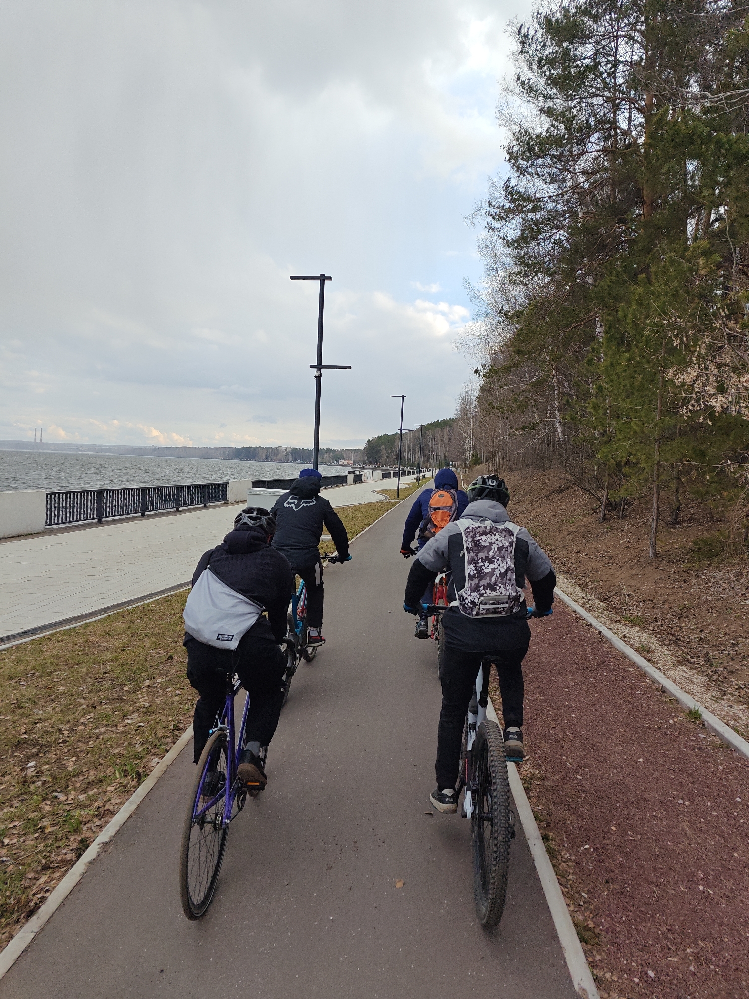 Four individuals are seen cycling from behind along a paved path next to a large body of water in Chaikovsky, Russia. The cyclists are wearing helmets, jackets, pants, and carrying backpacks. On the far left, one person rides a purple road bike and has a grey messenger-style bag. Next to them, another cyclist wears a black jacket with a white "FOX" logo. Two other cyclists follow on the right side of the path; one wears an orange backpack, and the last person has a white, grey, and black camouflage backpack, with "FILA" visible on their shoes.
The asphalt cycling path runs parallel to a concrete promenade lined with a black metal railing, separating it from a wide, choppy body of water, likely the Kama Reservoir. Across the water, distant land features and industrial structures, possibly power plant chimneys, are visible. To the right of the cycling path, there's a reddish gravel path alongside a dense forest containing a mix of evergreen and bare deciduous trees. The ground around the paths is covered with sparse grass and dry leaves, suggesting early spring or late autumn. Several modern black streetlights are spaced along the paths. The sky is largely overcast with some lighter patches. The scene depicts an outdoor recreational activity on a cloudy day.