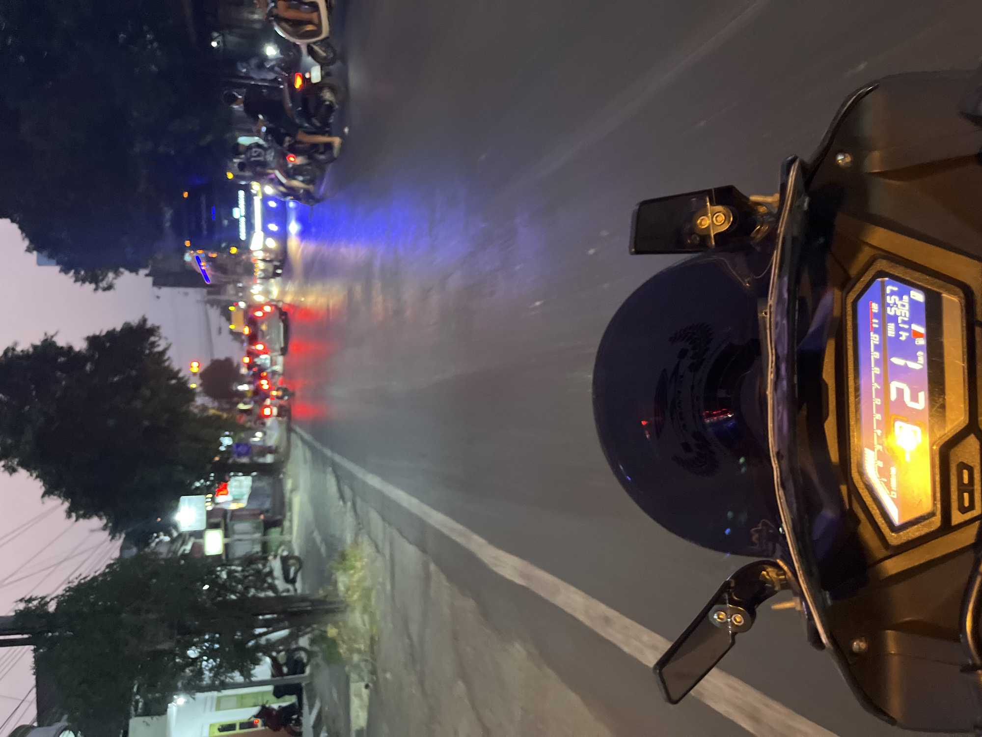 This image captures a view from a motorcycle in Kalinyamatan, Indonesia, at dusk. The rider is on a paved road with a white line marking the lane. To the left, a sidewalk with grass and some trees is visible, bordering a building. The road is moderately busy with traffic, including cars and motorcycles, illuminated by streetlights and the vehicles' headlights and taillights. A large truck with blue accent lighting is prominent in the distance. The motorcycle's digital dashboard is illuminated, displaying "5:57" and "41730". The sky is a pale twilight hue, and the overall atmosphere suggests a typical evening scene in a Southeast Asian city.