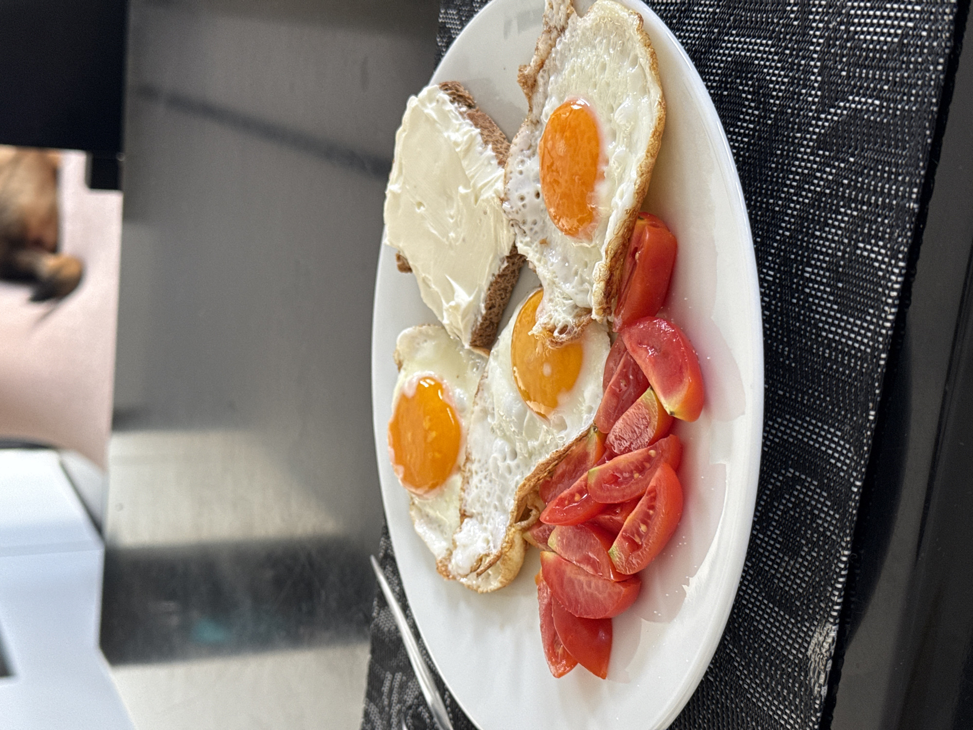 A white plate of food is centered in the frame, consisting of three fried eggs with bright orange yolks, sliced tomatoes, and a slice of dark bread topped with a generous layer of white cream cheese. The plate sits on a black and white woven placemat. To the left of the plate is a reflective surface and a partially visible animal, likely a cat, out of focus. The setting appears to be indoors, possibly a home kitchen or dining area. There is no information to determine if the picture was taken in Sofia, Bulgaria, and therefore the location is not specified in the description.