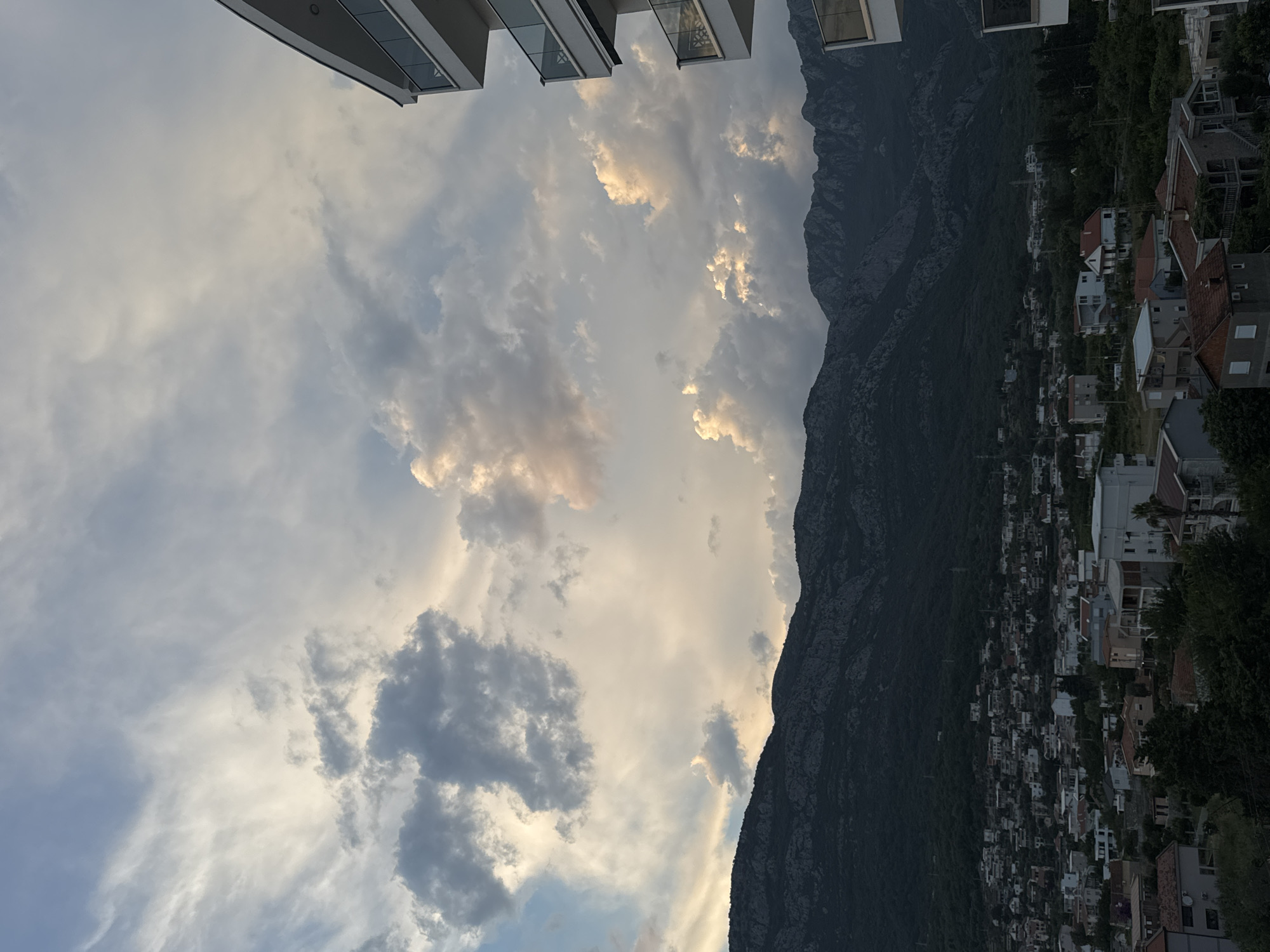 A wide shot captures the rugged, dark silhouette of a mountain range dominating the background, its slopes covered in dense green foliage. A coastal town, identified as Šušanj, Montenegro, sprawls across the foothills, a dense collection of buildings with terracotta roofs and white facades interspersed with trees. The sky above is a dramatic canvas of clouds, with patches of bright, illuminated white and soft pastels contrasting with darker, more ominous grey formations. To the far right of the frame, the modern white facade of a multi-story building with glass-paneled balconies is partially visible, extending upwards into the cloudy sky. The overall lighting suggests either early morning or late afternoon.
