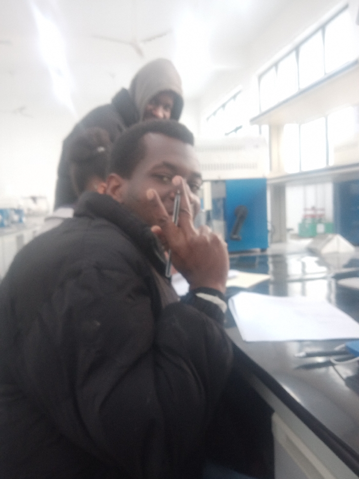 Two men are indoors. The man in front wears a black jacket and holds a pen in his right hand, gesturing a peace sign near his eye. A piece of paper sits on the dark surface in front of him. Behind him, another man is wearing a hooded jacket. Bright windows are visible in the background. There is equipment visible in the background, suggesting the location might be a laboratory.