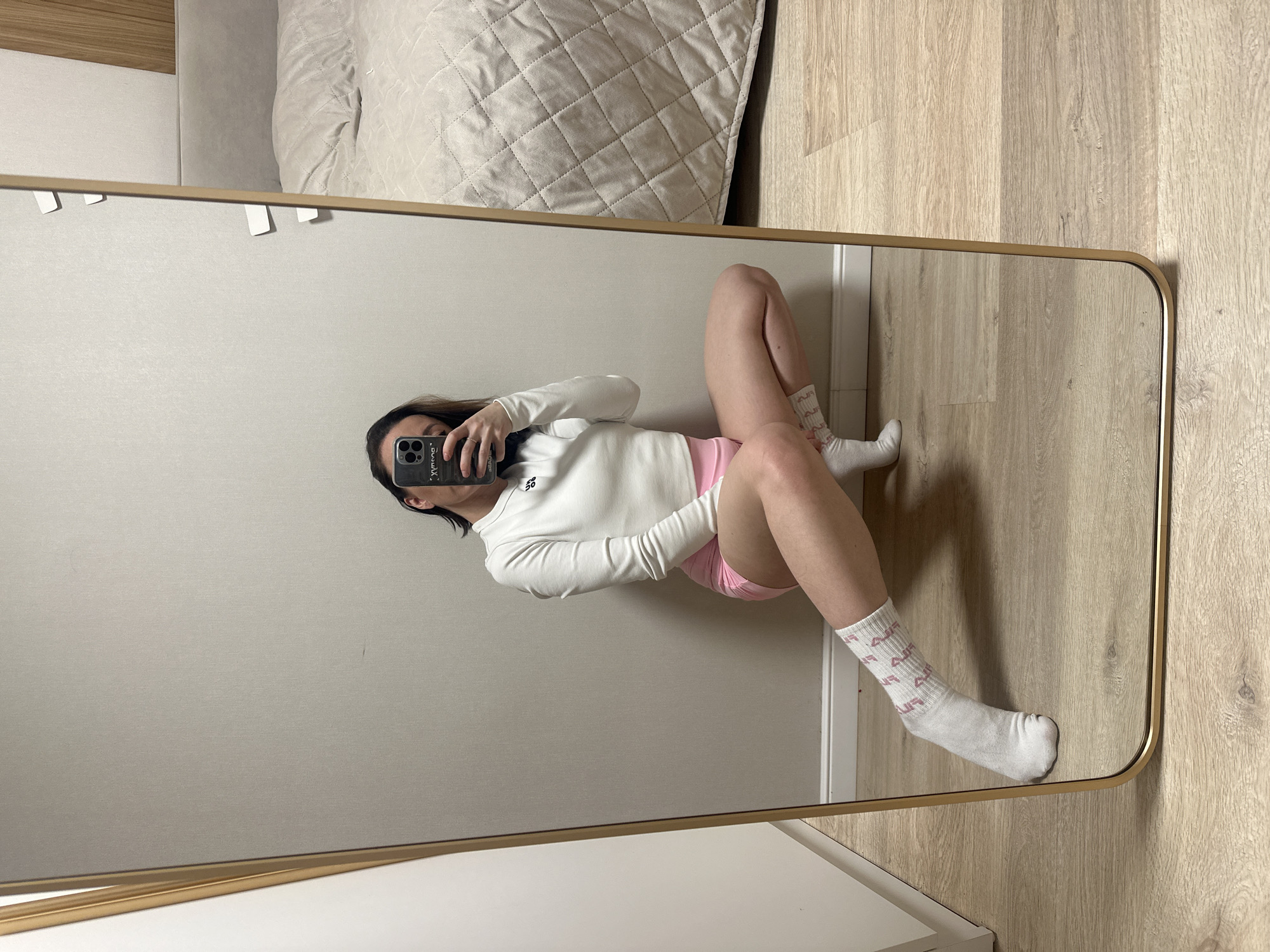 A mirrored selfie features a woman in a light-filled interior. She is positioned against a wall adjacent to a bed with a light-colored, quilted comforter. The woman is wearing a long-sleeved white shirt, pink shorts, and white socks with pink lettering. She holds a smartphone in her hand, presumably taking the picture. The reflection shows the room's interior, characterized by light-colored wood flooring and neutral wall paint. There are no distinctive elements visible to suggest that the location is within Votkinsk, Russia.