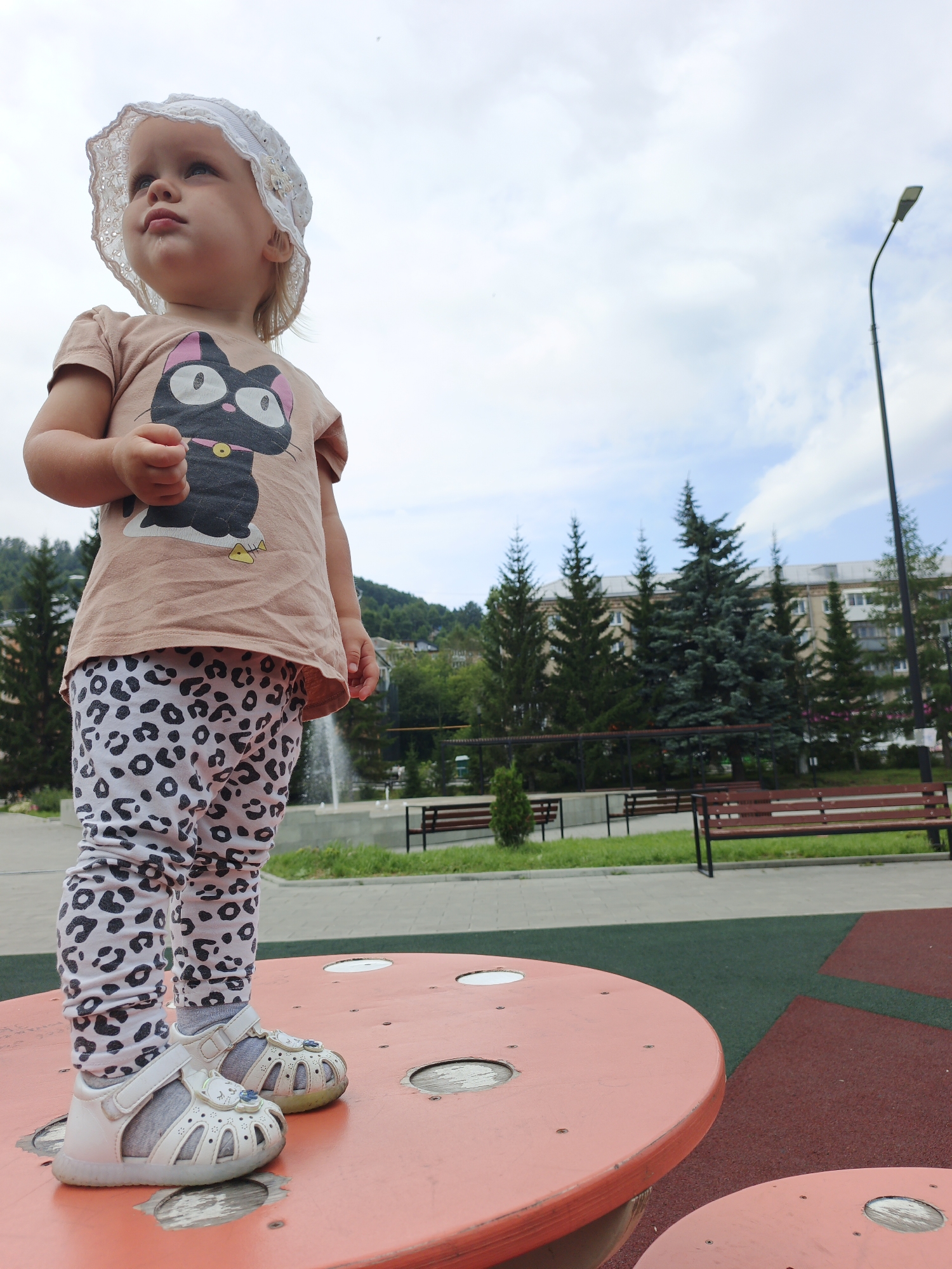 A low-angle, full-body shot captures a young Caucasian girl standing on a circular, orange-colored play structure in an outdoor playground. She is wearing a white, lacy sun hat, a tan t-shirt with a graphic of a black cat with pink ears and a yellow fish, and white leggings with a black leopard print pattern. Her white sandals have straps and decorative blue embellishments. The girl is looking upwards and to her left, with her right hand clenched. The playground features a rubberized surface with green and red sections. In the background, there are several dark green evergreen trees, a building with balconies, a street light, benches, a fountain, and a hilly landscape under a cloudy sky. This playground is likely located in Zlatoust, Russia, based on the architectural style of the building and the prevalent evergreen trees in the region.