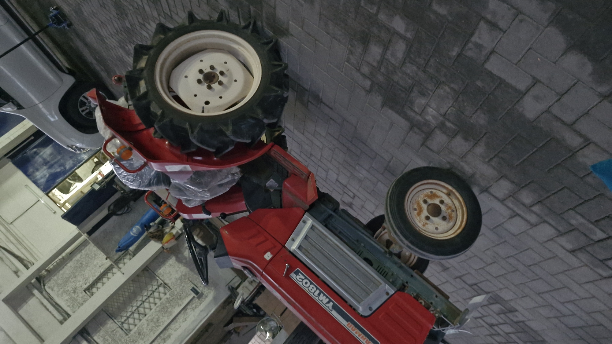 The image shows a red tractor in what appears to be a storage area or garage, with a white car in the background. The tractor is the primary subject, with its large, treaded rear tires and smaller front wheels prominently visible. The tractor has "YANMAR YM1802" written on its side in white lettering. It is partially covered in plastic sheeting, suggesting it may be new, stored, or being protected.
To the left of the tractor, a white car is partially visible, also covered. The ground is paved with interlocking bricks. Other objects in the scene include a blue cylinder or hose, various tools, and some shelving units on the left side, suggesting an organized but cluttered workspace. The lighting is artificial, indicating an indoor or covered location. The overall impression is one of a workshop or storage facility where vehicles are kept. There are no people visible in the image. The scene seems to be set during the day, but the lighting suggests it could be indoors or under artificial light. The location context provided is "Taraz city administration, Kazakhstan," which may hint at the type of facility this is, possibly related to municipal maintenance or agricultural equipment.