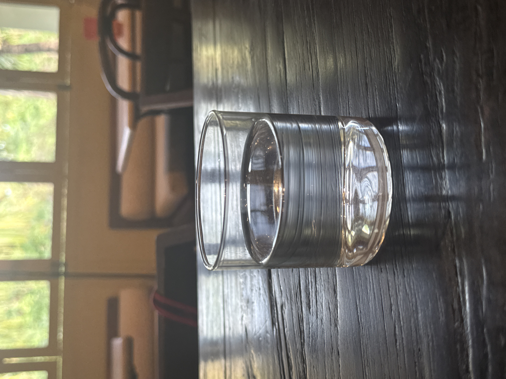 A clear, cylindrical, double-walled glass, partially filled with liquid, sits prominently on a dark wooden table surface. The table exhibits a distinct vertical wood grain pattern and reflects the ambient light, creating bright streaks.
The background is out of focus. To the left, a multi-paned window reveals lush green foliage and natural light from outside, suggesting an outdoor natural environment or garden. Further in the blurred background, elements of indoor furniture, possibly chairs with dark frames and lighter-toned upholstery, are faintly visible.
The scene is illuminated by bright natural light, indicating daytime, likely morning or afternoon, with sunlight casting reflections on the glass and table. There is no specific activity taking place, depicting a still life. No individuals are present, and no text is visible within the image. The setting suggests an indoor space with a view of nature, fitting a tropical context like North Kuta, Indonesia.