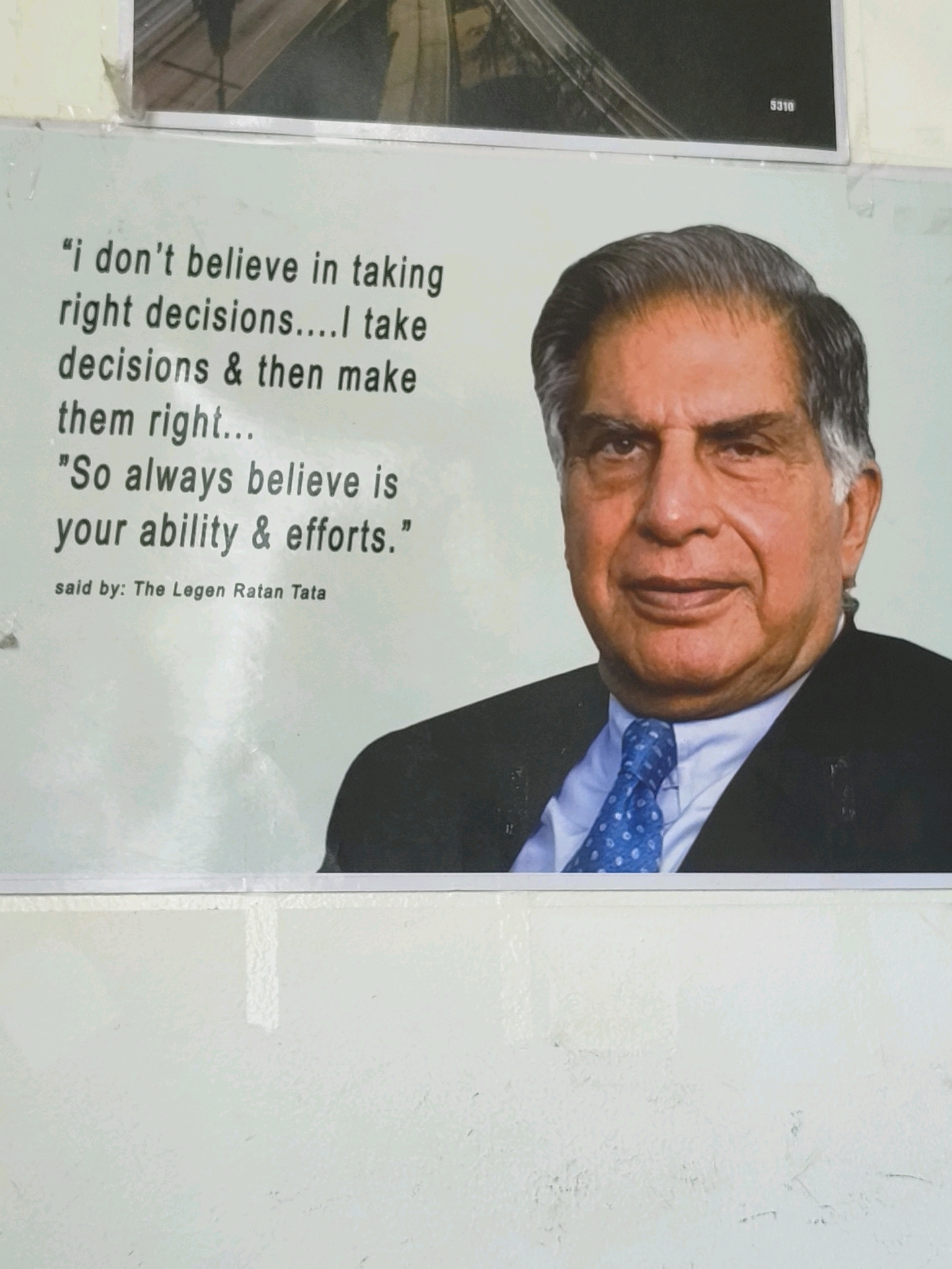 The image shows a framed motivational poster with a quote attributed to Ratan Tata, overlaid on a photograph of Ratan Tata himself. The quote reads: "i don't believe in taking right decisions....I take decisions & then make them right... 'So always believe is your ability & efforts.'" Below the quote, it states, "said by: The Legen Ratan Tata". The photograph depicts Ratan Tata wearing a suit and a blue polka-dot tie. The poster is mounted on a wall, and the bottom portion of the image reveals a textured, off-white surface, likely a painted wall. There is a subtle hint of a white vertical line or seam in the wall behind the poster. The top portion of the image shows a blurry black and white photograph, possibly of an infrastructure like a bridge or highway, with the number "5310" visible in the top right corner of that photograph. Based on the provided context that this image is located in Tathawade, India, this poster is likely displayed in a public space, office, or educational institution within Tathawade.