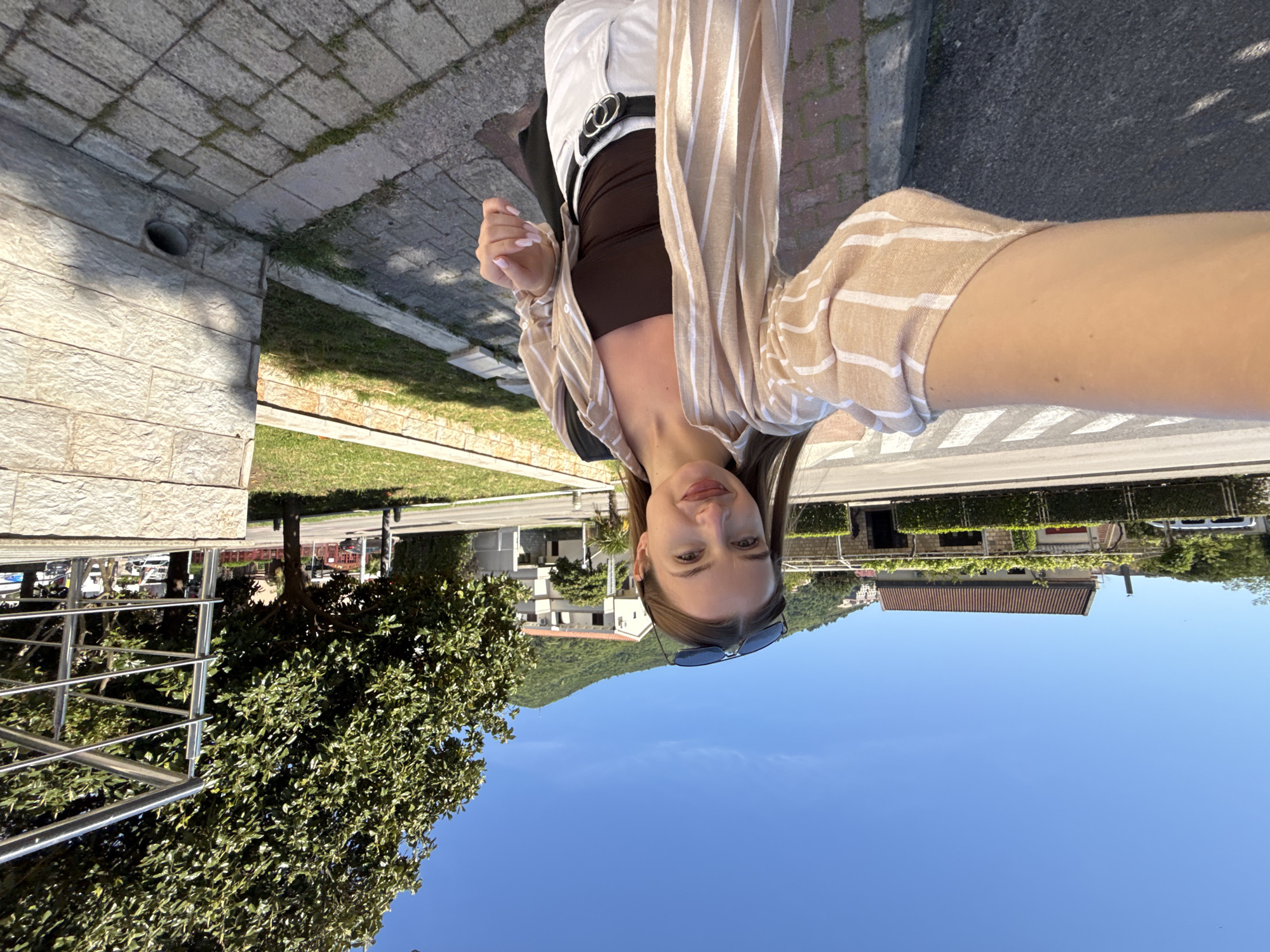The image is a bright, outdoor selfie taken by a young woman in Budva, Montenegro. The scene is set on what appears to be a sidewalk or paved area, with a stone wall and green grass to the left, and a road with buildings and lush hills in the background.
The primary subject is a young woman, captured from the chest up, looking directly at the camera with a slight smile. She has light brown hair, which is styled back, and a pair of dark sunglasses rests on top of her head. She is wearing a casual but stylish outfit consisting of a dark brown fitted top or camisole, white trousers or pants, and a loose-fitting, unbuttoned striped shirt. The shirt features beige or light tan and white vertical stripes. A black belt with a prominent circular silver buckle cinches her waist. Her fingernails are painted a light, neutral pink shade.
The background shows a clear, bright blue sky dominating the upper portion of the frame. In the distance, rolling green hills or mountains are visible, characteristic of a coastal European landscape. Closer to the foreground, on the opposite side of the road, are several buildings with light-colored walls and traditional red-tiled roofs. Green trees and other foliage are scattered throughout the scene. A silver railing, possibly part of a fence or balcony, is visible on the left side of the image.
The image was taken during bright daylight, suggesting a sunny day with clear weather. There is no visible text in the image. The overall impression is one of a relaxed, pleasant moment in a scenic location.