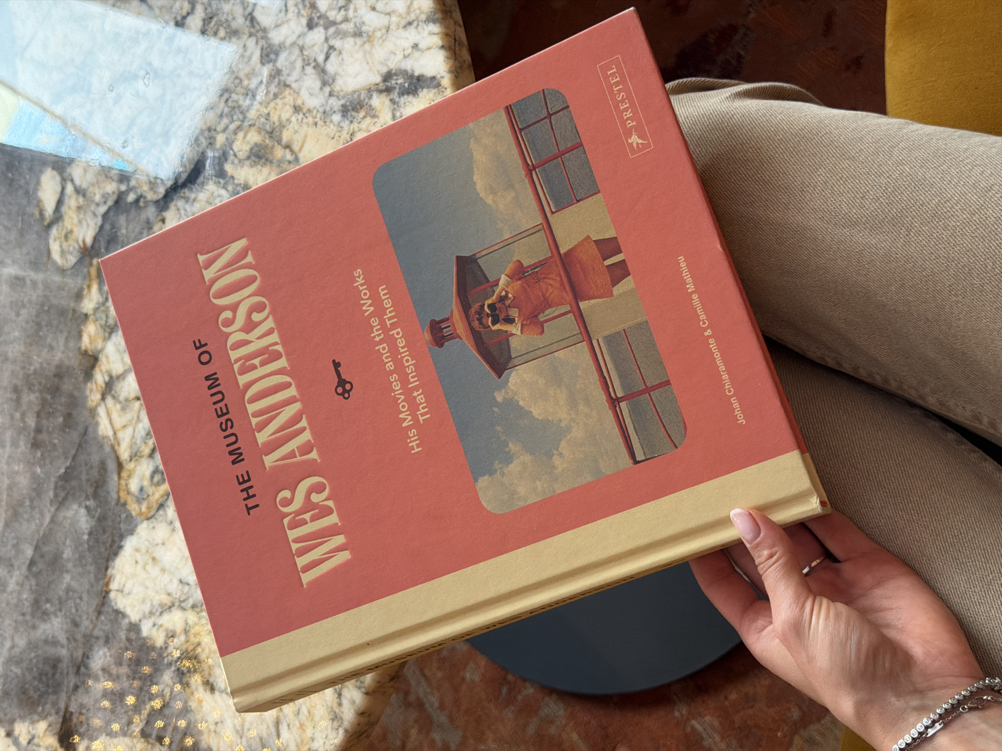 A woman's hand with a silver ring and bracelet is holding a book titled "The Museum of Wes Anderson: His Movies and the Works That Inspired Them." The book has a coral-colored cover with gold lettering. A small image on the cover depicts a woman in an orange dress looking through binoculars in a tower. The book is lying on a marble countertop, and part of a beige fabric garment is visible. The background shows a blurred environment with a dark blue circular object and what appears to be a yellow upholstered chair. No discernible elements indicate the location in Saint Petersburg, Russia.