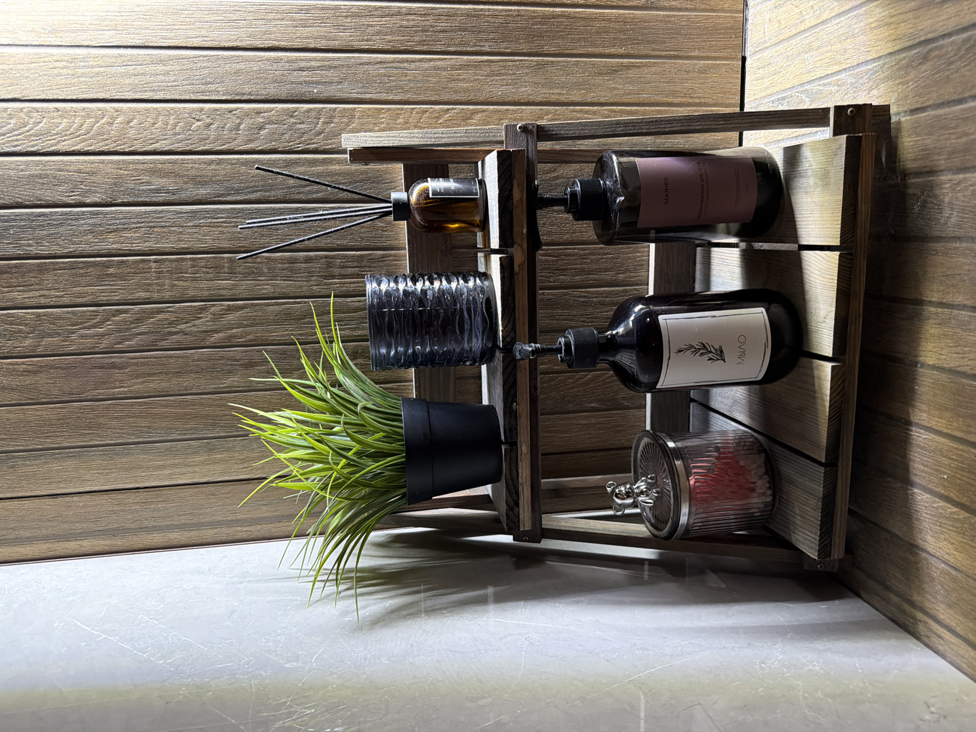 The image depicts a two-tiered wooden shelf against a backdrop of vertical wooden planks. On the top shelf, from left to right, are a black potted plant with green, spiky leaves, a dark, ribbed glass container, and a small amber glass bottle with a diffuser stick. On the bottom shelf, also from left to right, are a clear glass jar with a silver lid topped with a small bear figurine and containing pinkish objects, a dark bottle with a pump dispenser and a white label, and another dark bottle with a pump dispenser and a pink label. The lighting suggests an indoor setting, possibly a bathroom or vanity area. The overall scene is static and appears to be a still life arrangement. There is no indication of the city of Semiluki, Russia, or any specific location within it.