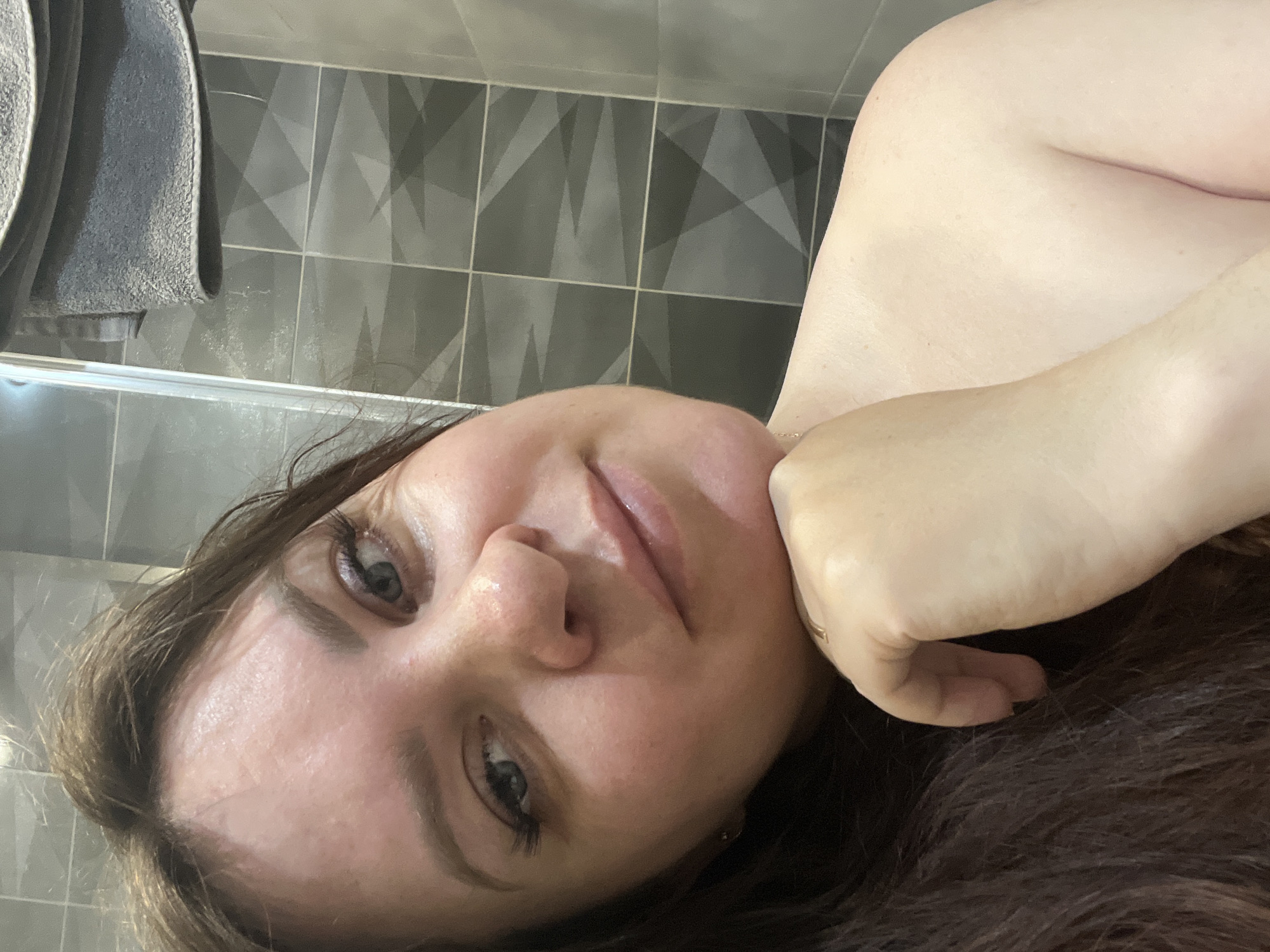 A close-up, angled self-portrait of a woman with fair skin and brown hair. The woman's head rests in her hand. Her eyes are open and appear to be light-colored. The background features gray tiles with a geometric pattern and a gray towel rack. The location is indeterminable from the provided image. The city of Noviy, Russia is not discernible from the image.