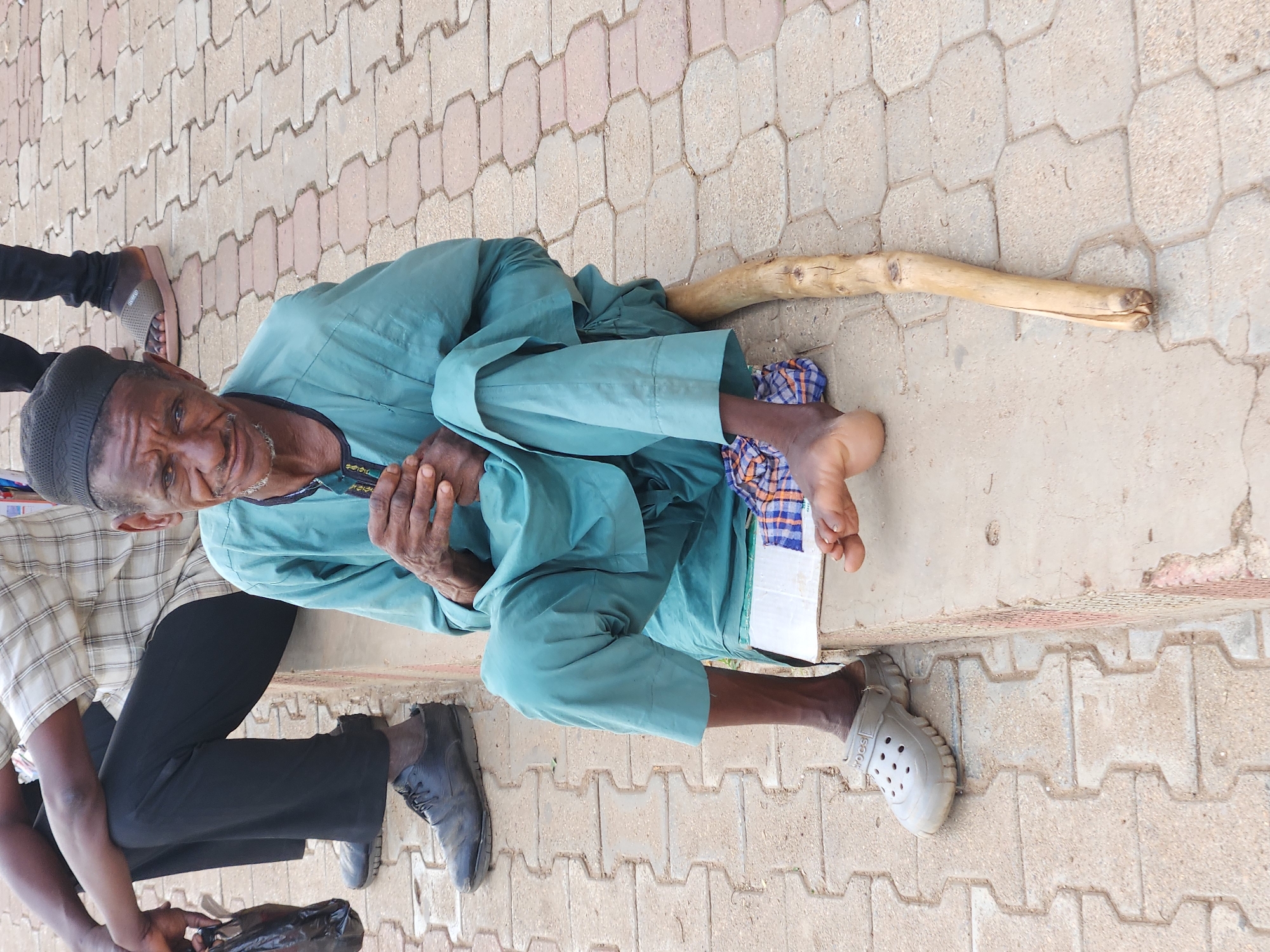 A medium-angle shot depicts an elderly man in light blue clothing sitting on a patterned brick sidewalk in an urban environment. The man has dark skin, a white beard, and a black hat. He is holding his hands together, and his face is visible with a slight smile. Next to him is a wooden stick, and he is wearing one grey Croc-like shoe.
To the left of the elderly man, portions of two other individuals are visible. The closest person wears a light plaid shirt and holds a black bag. The third person wears black pants and brown sandals.
The sidewalk is comprised of interlocking bricks of various light tones. The background is out of focus, and the specific city and location are unclear.