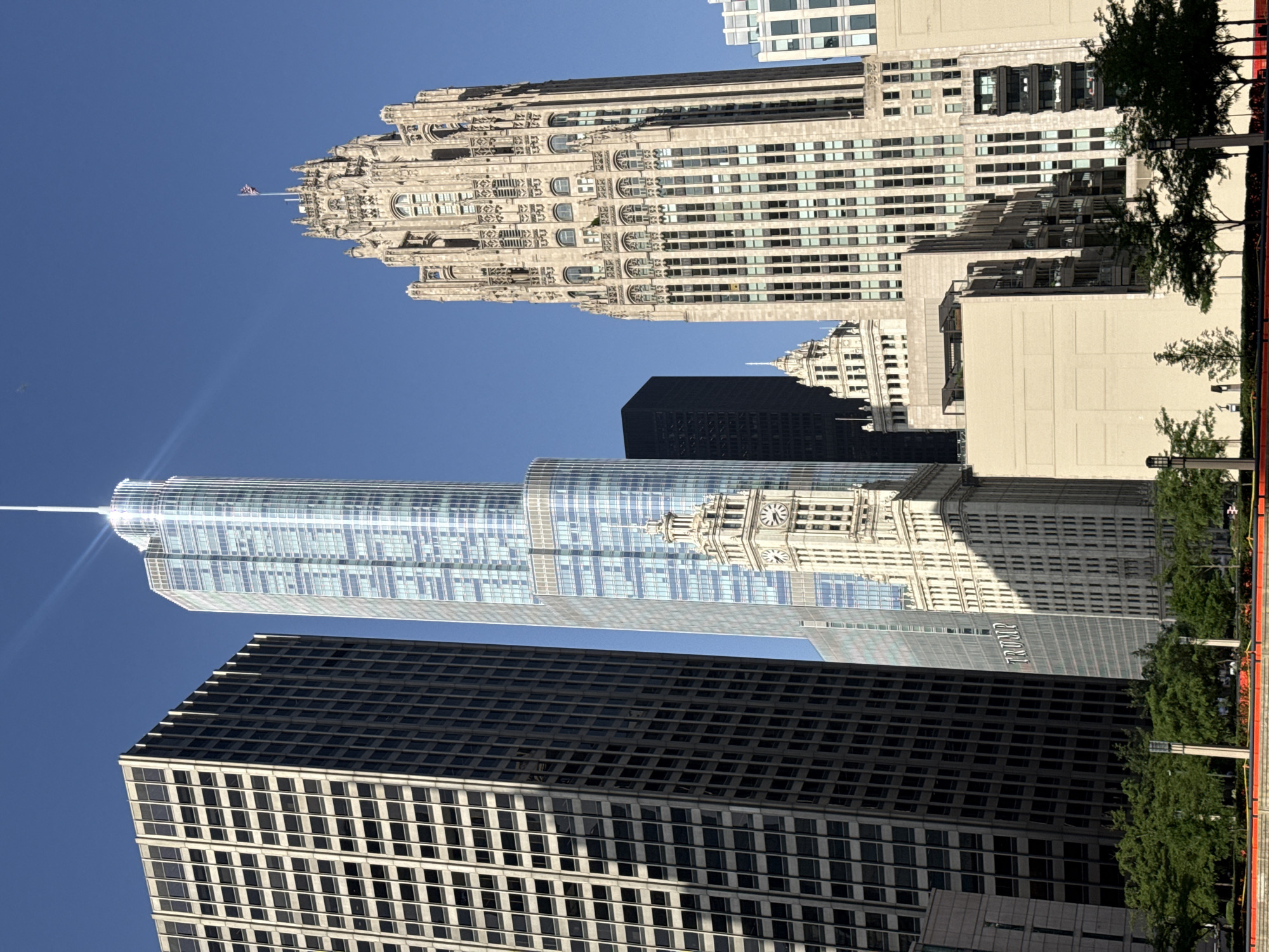 This media file captures a view of several skyscrapers in Chicago, United States of America. The scene is dominated by towering buildings with distinct architectural styles. In the background, a light-colored, ornate skyscraper with gothic-inspired detailing stands prominently against a clear blue sky, suggesting a bright, sunny day. In the mid-ground, a modern glass skyscraper with a reflective facade reflects the sunlight, appearing almost ethereal. To its right, a building with a large clock face is visible, hinting at a historic structure. In the foreground, a dark-colored, rectangular skyscraper with a grid-like pattern of windows occupies a significant portion of the frame, contrasting with the lighter buildings. The presence of green trees and some red-orange barriers at the bottom edge of the image suggests an urban outdoor setting, possibly a park or pedestrian area. The clear blue sky and bright sunlight indicate it is daytime, likely late morning or early afternoon. There is no visible text, people, or any active event taking place; the focus is solely on the architectural landscape of the city.
