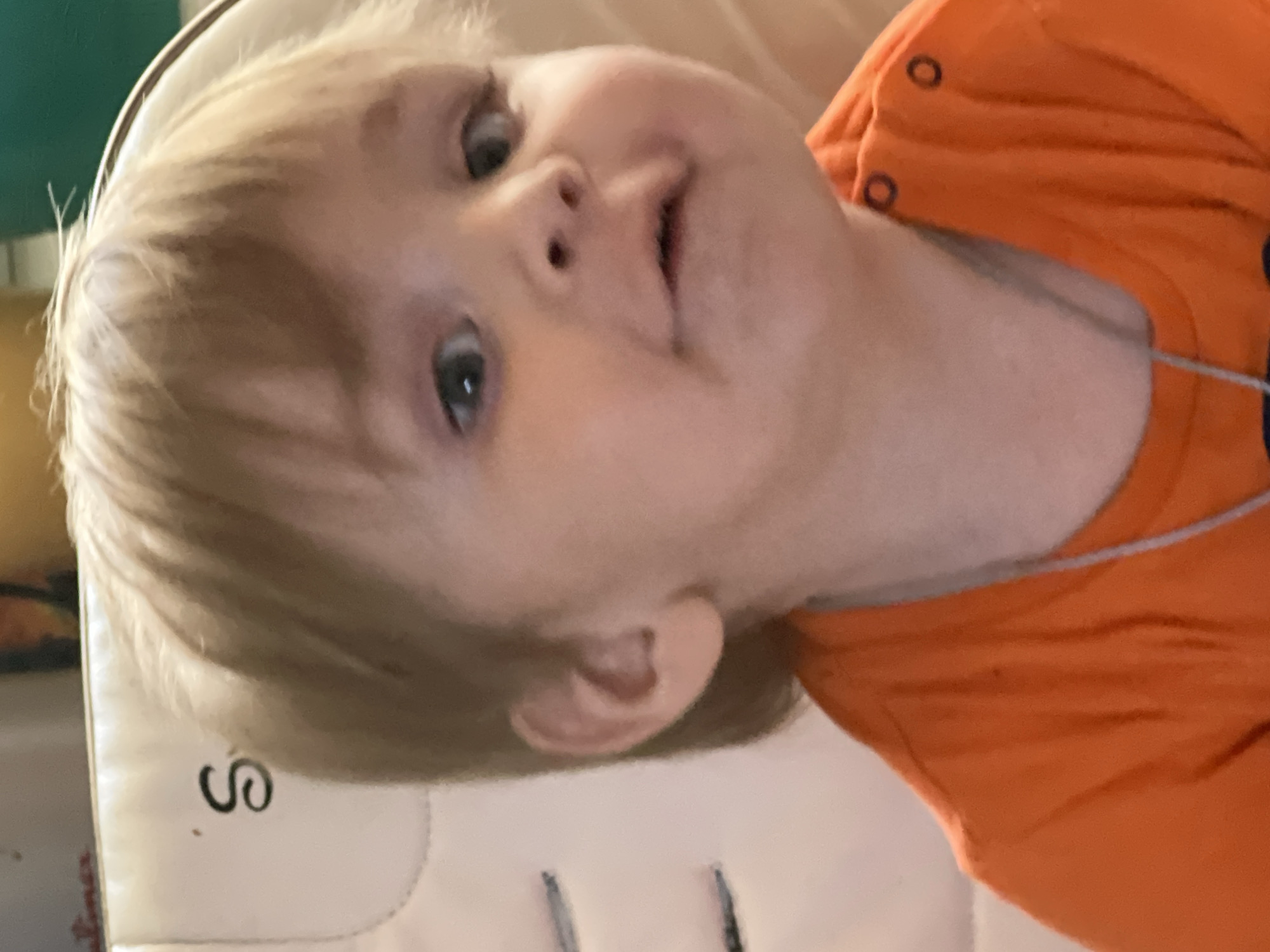 The image is a close-up portrait of a young child, likely a toddler, with blond hair and fair skin. The child is looking slightly to their right with wide, curious eyes and a slightly pursed mouth. They are wearing an orange t-shirt. The child appears to be seated in a white chair with a black letter "S" visible on its side. The background is blurred but suggests an indoor setting. There is no information in the image to indicate a specific location in the city of Noviy, Russia.