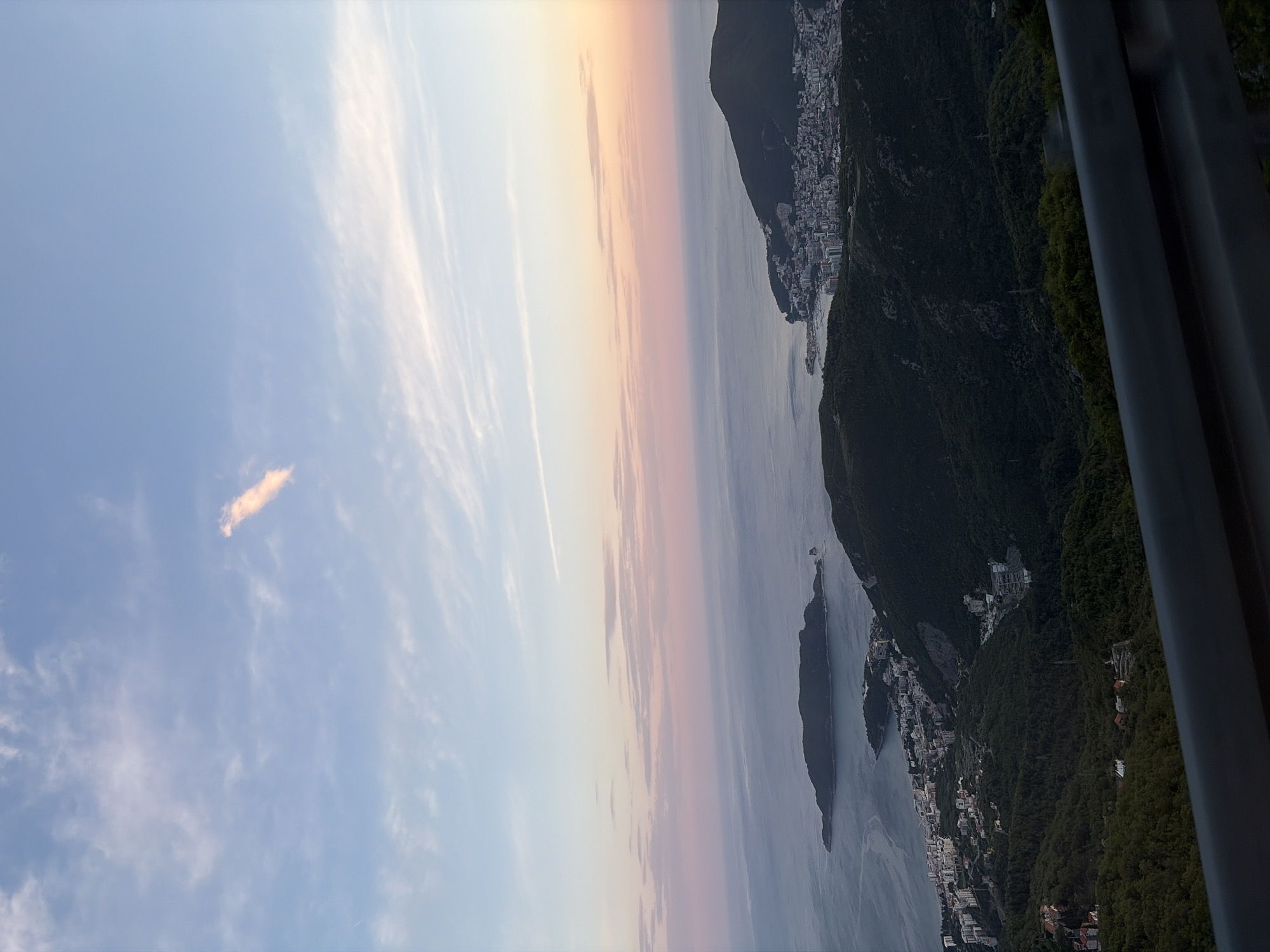 A landscape photograph displays the town of Brajici, Montenegro, from an elevated vantage point, possibly a cable car. The scene is divided into thirds. The left third shows a pale blue sky with wispy, light-colored clouds. The middle third is composed of a gradient of colors - light blue at the top blending into orange, pink, and eventually into the gray-blue of the sea. The right third is dominated by a dark green, mountainous landscape dotted with white buildings that appear to be residences or commercial structures. In the foreground on the far right are dark, blurred structures, potentially part of the infrastructure of the viewing platform.