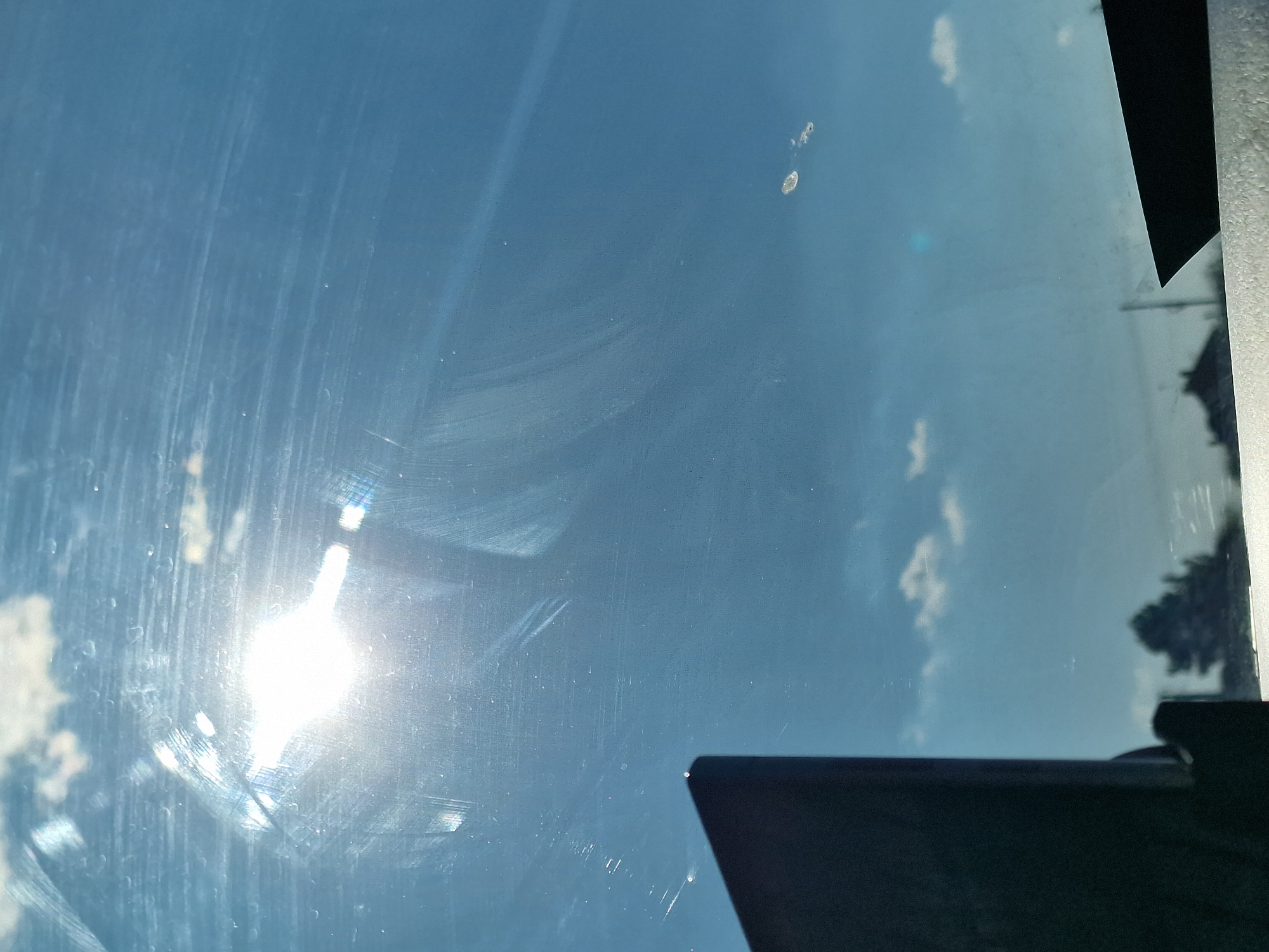 The image displays a bright, sunny daytime scene viewed through a dirty glass surface, likely a car windshield or window. The sky is a clear blue with scattered white cumulus clouds visible in the background. A prominent sunburst and associated lens flare dominate the lower-left section of the frame (when viewed with the sky horizontally), indicating direct sunlight.
The glass itself is heavily marked with numerous vertical and circular streaks, smudges, and what appear to be dried water spots or cleaning residues. In the upper-central area, there are a couple of distinct, light-colored marks, possibly dried bird droppings or similar debris.
Dark, angular structures intrude into the frame from the upper-right and lower-right corners. These appear to be parts of a vehicle's interior, such as a dashboard, window frame, or pillar. A faint reflection of dark foliage, resembling a tree or distant treeline, is also visible on the glass near the right edge of the frame.
The image captures a moment on a bright, fair-weather day, observed from inside a confined space looking outwards. No people are visible, and there is no discernible text. The overall scene suggests a need for window cleaning.