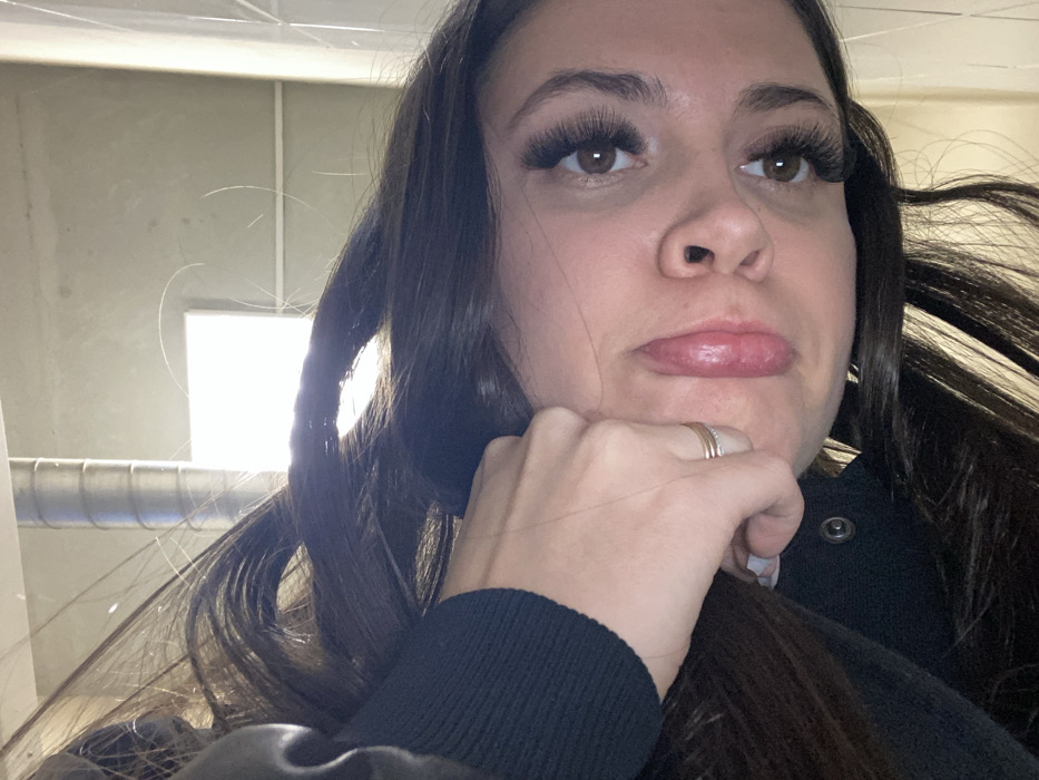 The image, rotated 90 degrees counter-clockwise from an upright perspective, depicts a close-up of a female subject in an indoor setting within the city of Chaikovsky, Russia. A young adult female with long, dark brown hair occupies the foreground, looking directly at the camera. Her left hand is positioned against the left side of her face/jawline, with fingers slightly curled. A ring, consisting of two thin, parallel gold-colored bands, is visible on her left ring finger. She wears a dark, possibly black, long-sleeved garment, appearing ribbed on the sleeve and featuring a single visible metallic snap button near the collar area. Her facial features include brown eyes with prominent, dark eyelash extensions, a discernible nose, and full, pink lips. The background consists of a light-toned wall or ceiling. A bright, rectangular light source, likely a window or light panel, is present in the upper background, casting a visible glare. A segment of a metallic, corrugated cylindrical duct or pipe is also visible in the upper left portion of the background.