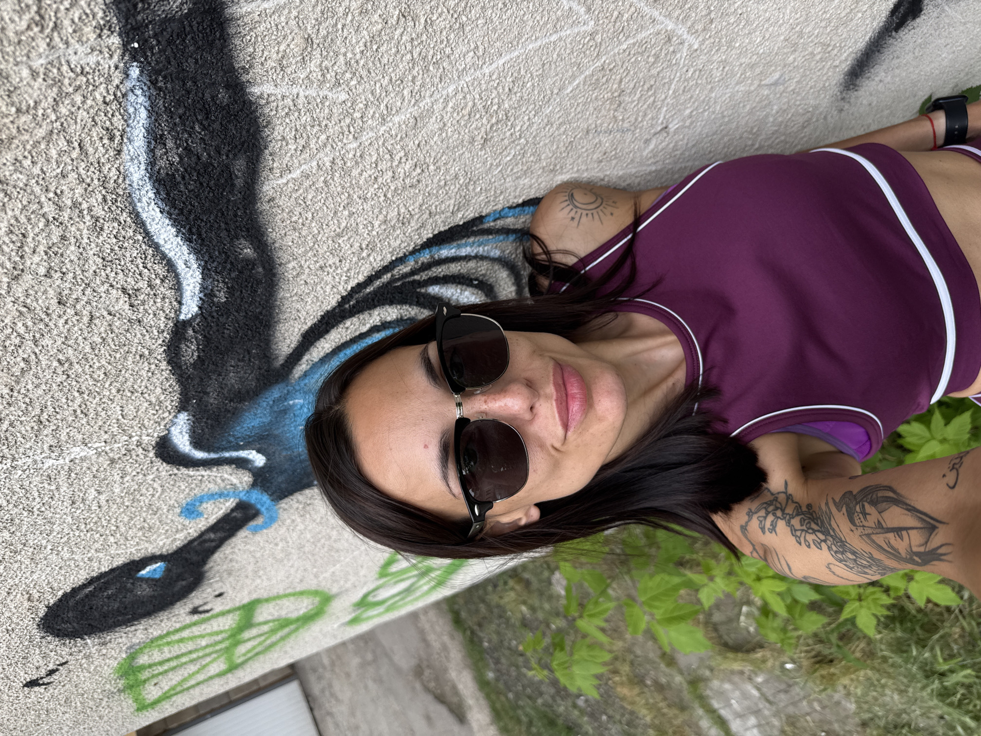 A woman wearing sunglasses and a plum-colored athletic top with white trim leans against a gray concrete wall covered in graffiti. The graffiti includes black and blue abstract shapes, as well as some green leaf-like shapes. The woman has dark hair, fair skin, and tattoos on her arms and shoulder. She is wearing a watch. Green plants grow near the base of the wall. The image appears to be taken at an angle, so the background is slightly blurred. While a forensic description can be provided based solely on visual content, information about the location in the city of Pleven, Bulgaria, cannot be determined without further data points beyond the provided image.