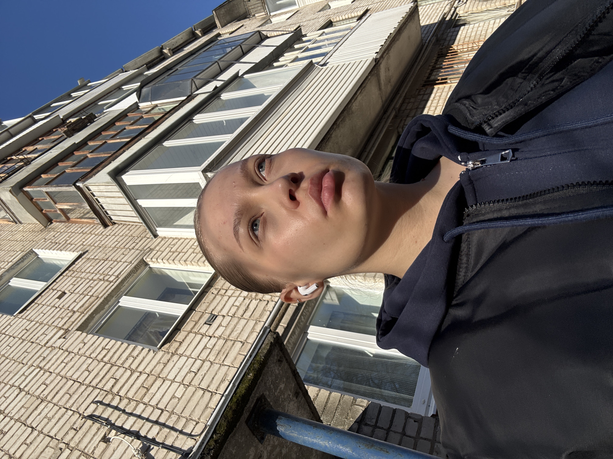 A medium shot depicts a fair-skinned person with a shaved head wearing a black jacket with a navy blue hood, looking upward with a slight frown. A white earbud is visible in their left ear. The background reveals a multi-story building with a mix of white-framed windows and brick facade. The image is tilted at an angle, suggesting the camera was positioned from below, looking up towards the subject and the building. A blue-painted railing is visible in the lower left corner. The scene is brightly lit by sunlight, casting shadows on the building's facade. The location appears to be in an urban setting, possibly in Chaikovsky, Russia, given the architecture and general environment.