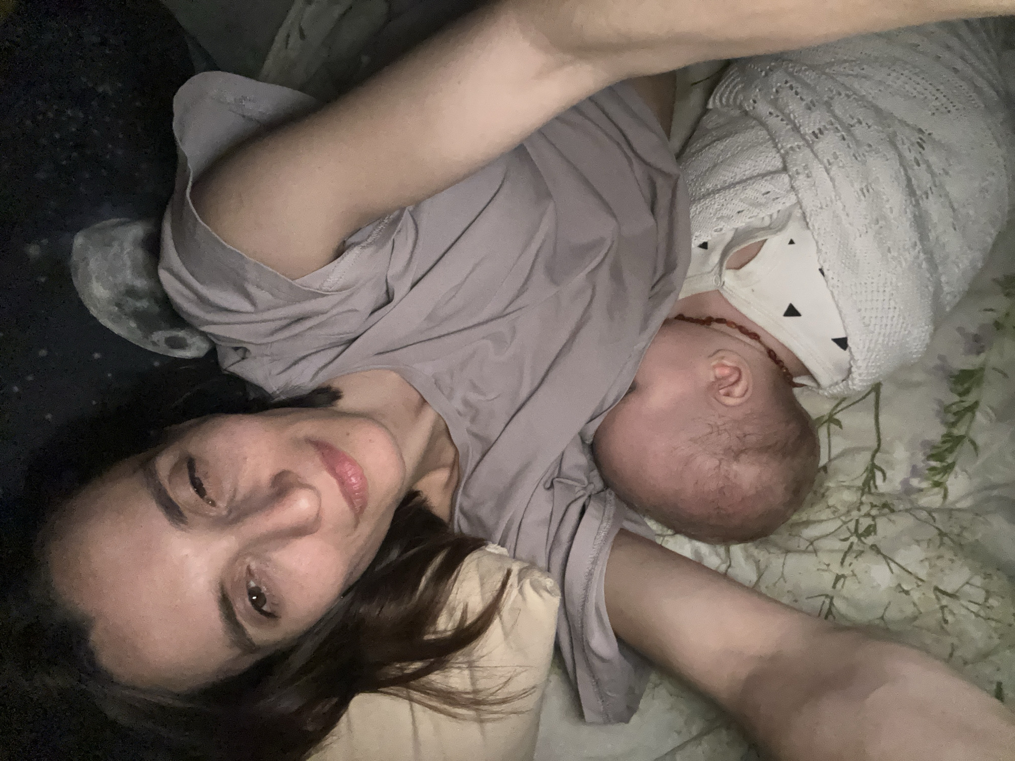 A woman with dark hair lies on a bed with a baby next to her. The woman is smiling and looking at the camera. The baby is wrapped in a white blanket. The bed has a floral pattern. The location cannot be determined from the image.