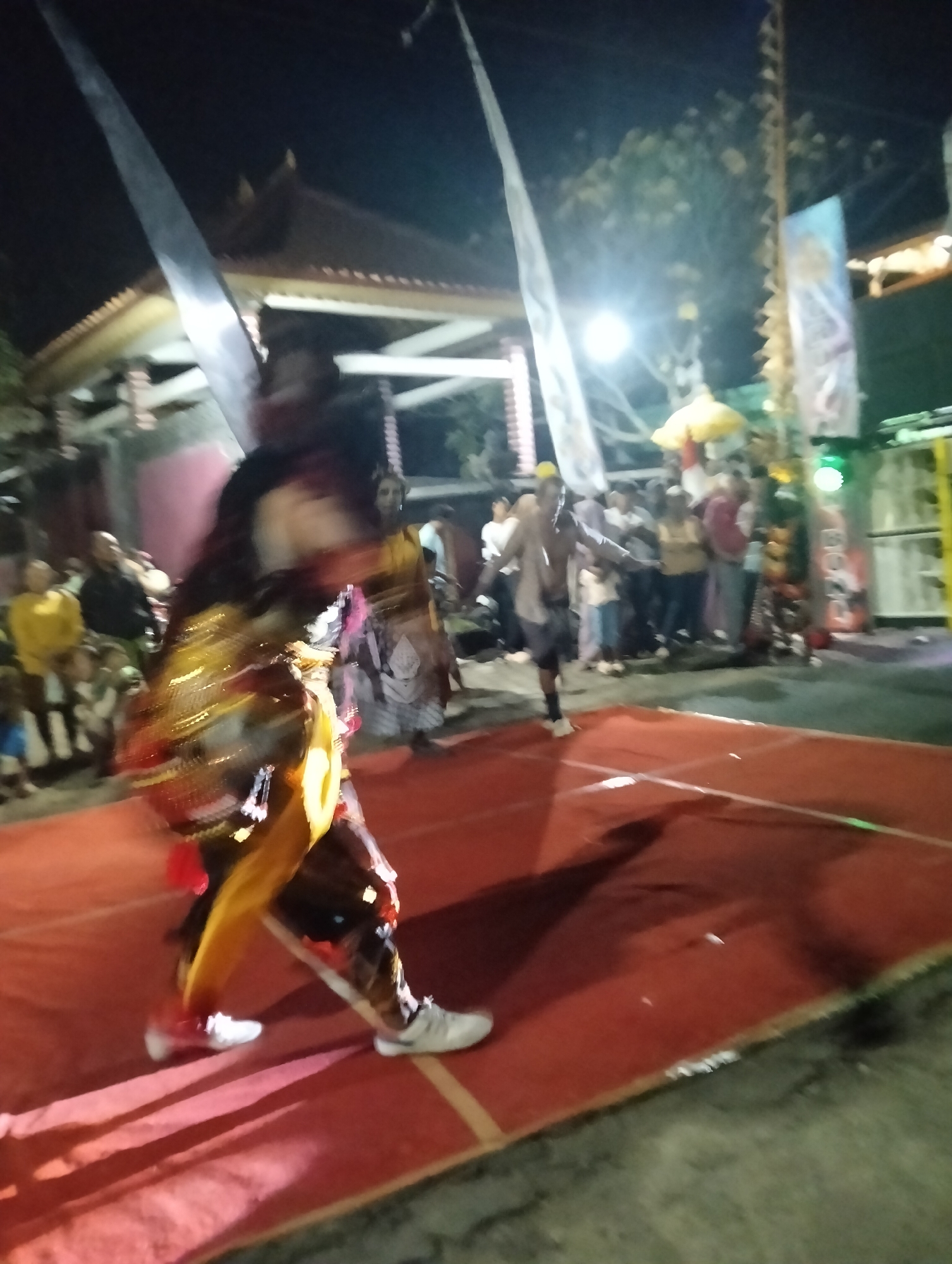 In Sumbersewu, Indonesia, at night, a performance takes place on a red stage. A dancer, in a gold and black costume with flowing yellow fabric, is the central figure, captured in motion. The dancer's face is blurred, indicating movement. A crowd of onlookers is gathered in the background, partially obscured by the darkness and distance. Some individuals are wearing casual clothing, while others appear to be in traditional attire. Tall, slender flags or banners are visible behind the stage, adding to the festive atmosphere. The environment suggests an outdoor event, possibly a cultural festival or celebration.