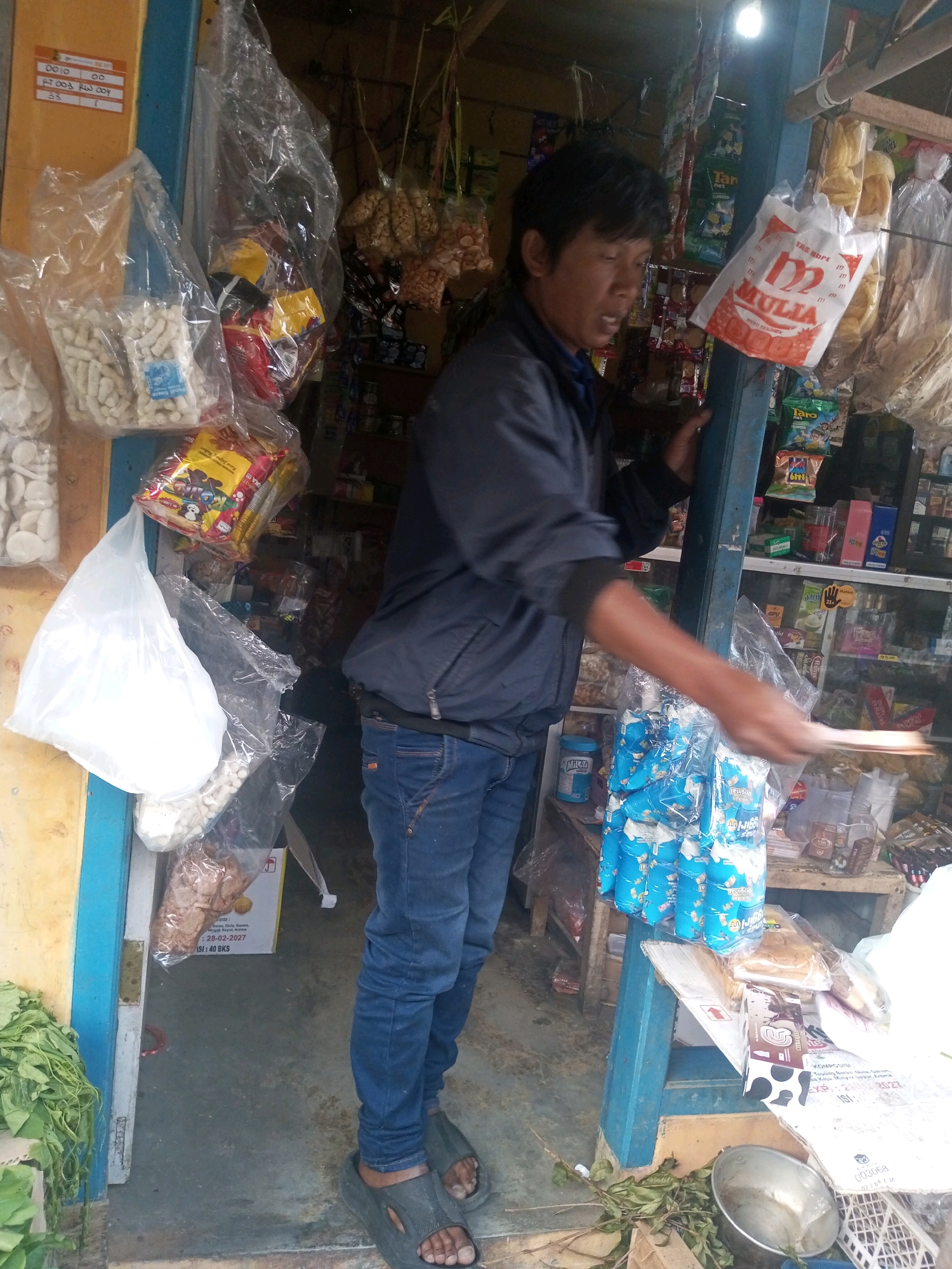 A man stands in front of a small shop in Andongsili, Indonesia, with his arm extended, holding a piece of paper. He is wearing a dark jacket and blue jeans. The shop is filled with various goods, including bags of snacks, drinks, and other items. The shelves behind him are stocked with more products. In the foreground, there is a pile of green leaves and a metal bowl. The shop appears to be open for business.