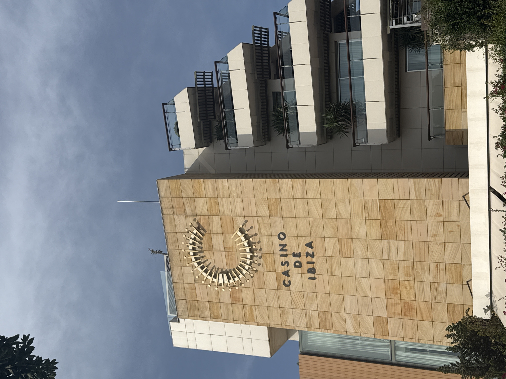 The image shows the exterior of the Casino de Ibiza building, located in Eivissa, Spain. The building has a modern architectural design with a facade clad in light-colored stone tiles. A prominent circular logo, resembling a stylized 'C', is affixed to the stone facade, and the text "CASINO DE IBIZA" is displayed below it in dark, sans-serif lettering. The building features a series of staggered balconies with glass railings and wooden accents, giving it a tiered appearance. Greenery, including small trees and plants, is visible on some of the balconies and at the base of the building. The sky above is partly cloudy. There are no people visible in the image.