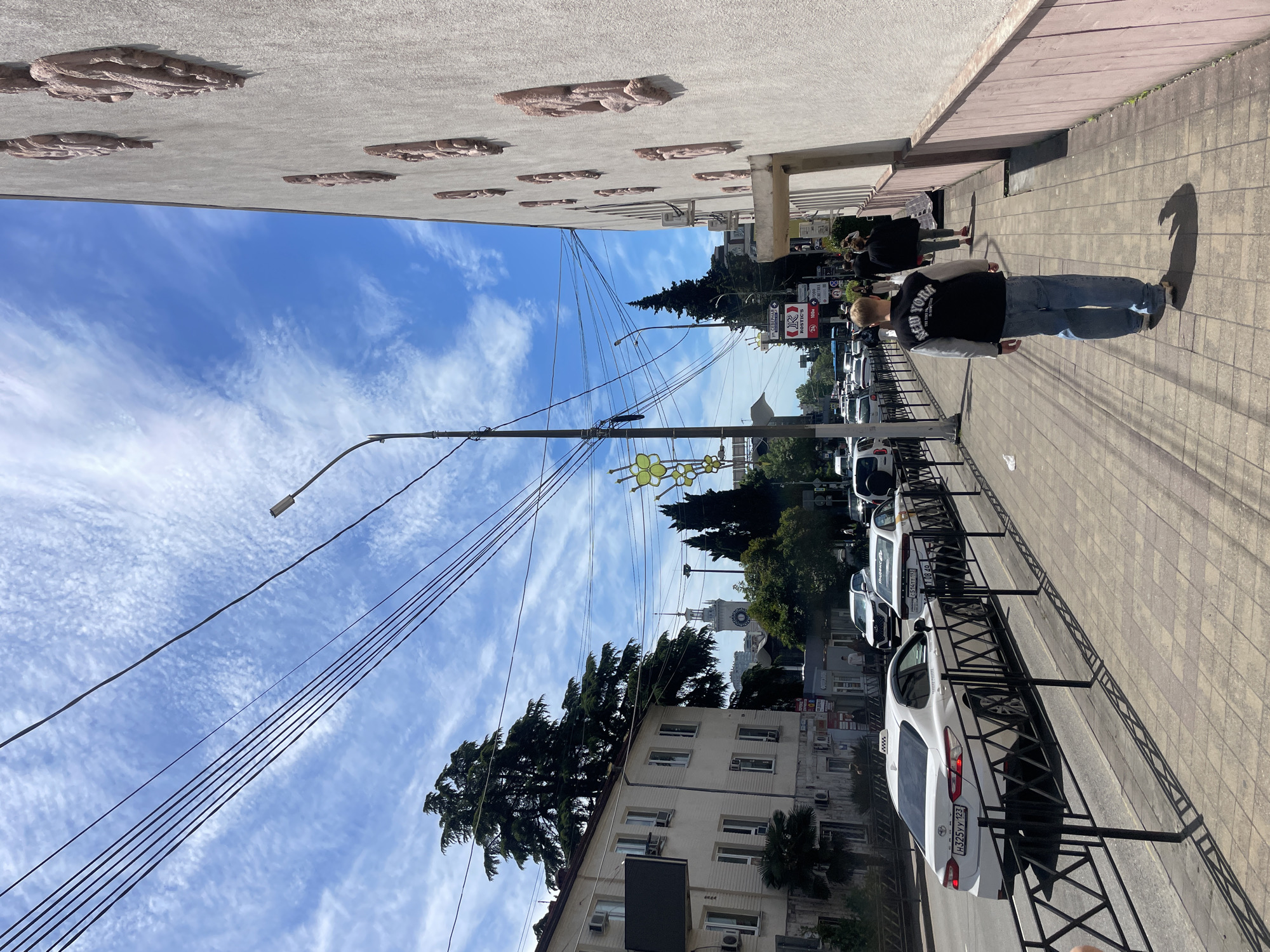 A street scene in Sochi, Russia, shows a man walking along a sidewalk. He is wearing a black jacket with "NEW YORK" printed on the back and blue jeans. A row of parked cars lines the street, separated from the sidewalk by a low black metal fence. Above the cars, a building with white walls and multiple windows stands. Tall green trees punctuate the street scene. Overhead, power lines crisscross against a sky filled with wispy clouds. In the distance, the spire of a building is visible. The sidewalk is paved with rectangular tiles. The wall of the building next to the sidewalk is decorated with a series of raised, textured elements. A streetlight and decorative floral light fixtures are also visible.