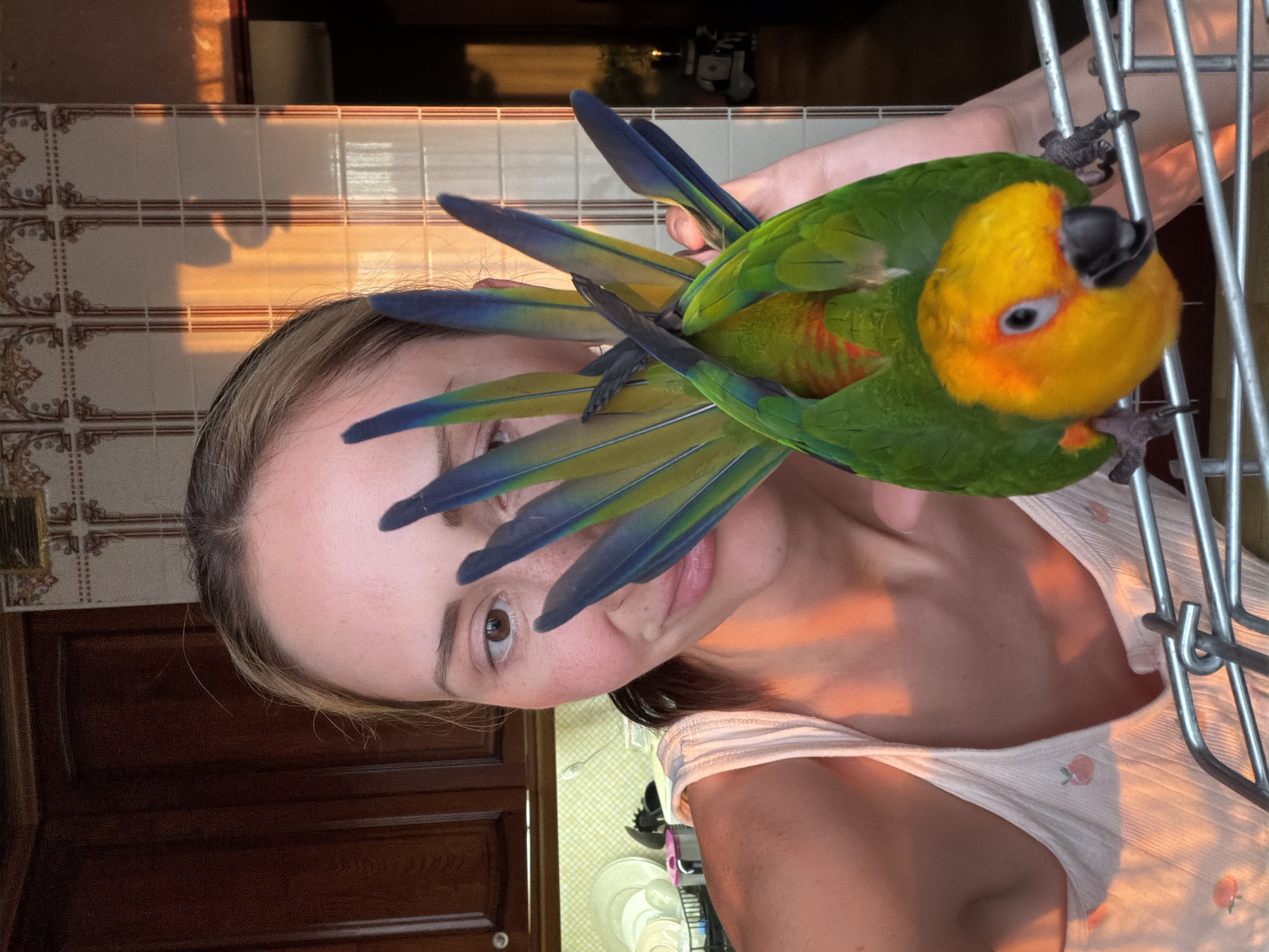 A woman is holding a green and yellow parrot. The parrot is perched on a metal cage. The woman's face is partially obscured by the parrot's tail feathers. The background includes a tiled wall and a wooden door. The location is not discernible as Khimki, Russia based on the provided visual information.