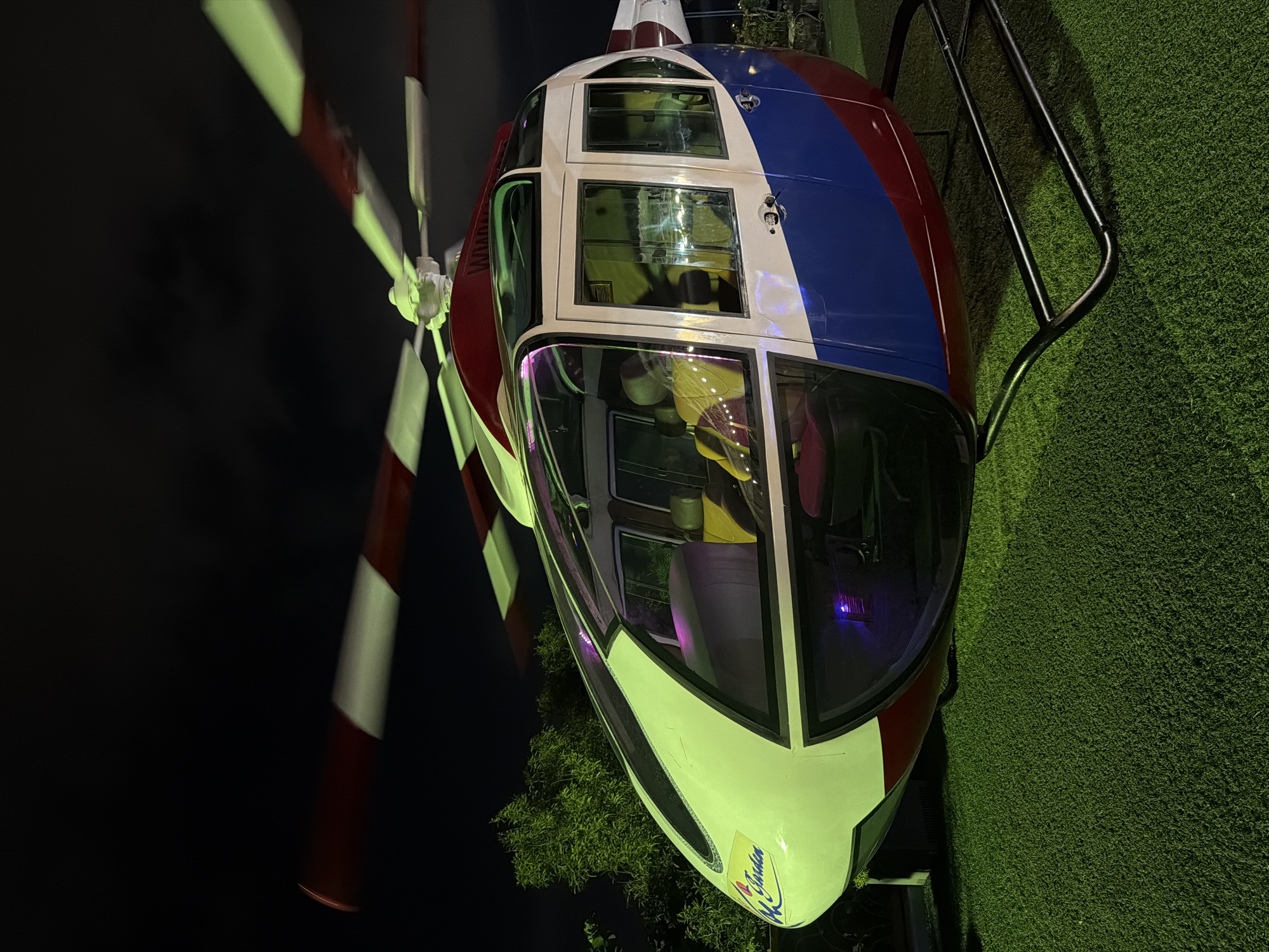An outdoor nighttime photograph captures a helicopter that appears to be a static attraction on artificial turf. The helicopter is predominantly white with red and blue accents along its fuselage. Its rotor blades, a mix of red and white, are blurred, suggesting motion, though the overall impression is of a stationary display. Inside the cockpit, a person wearing a yellow shirt is visible. The environment is dark, with the helicopter illuminated by artificial lights that cast a greenish glow on the turf and a purple light within the helicopter's cabin. The background is dimly lit, with the silhouettes of trees visible to the left. The helicopter is mounted on a black metal stand. A sign on the front of the helicopter reads "A Garden." The location is specified as Nam Nha Trang Ward, Vietnam.